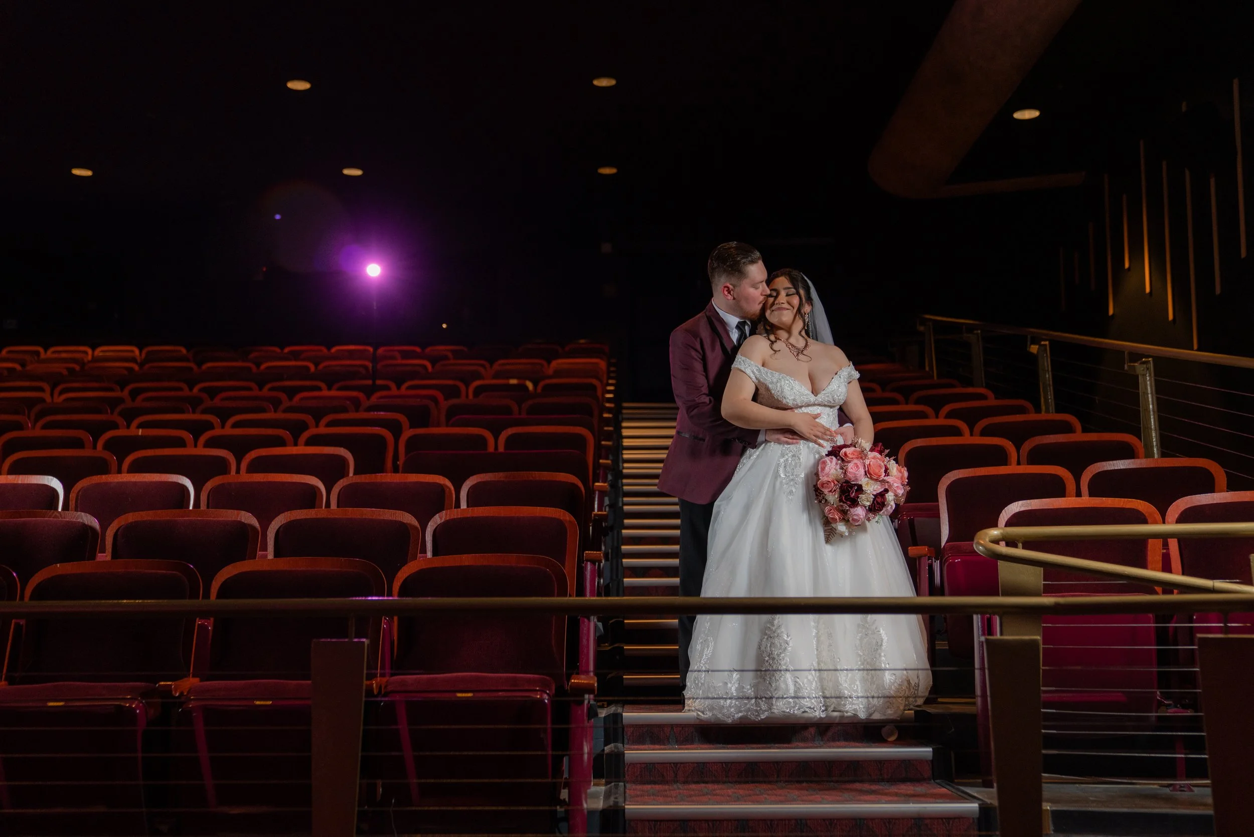 A bride and groom share a kiss in an empty theater auditorium with red seats and dim lighting, with a pink stage light in the background.
