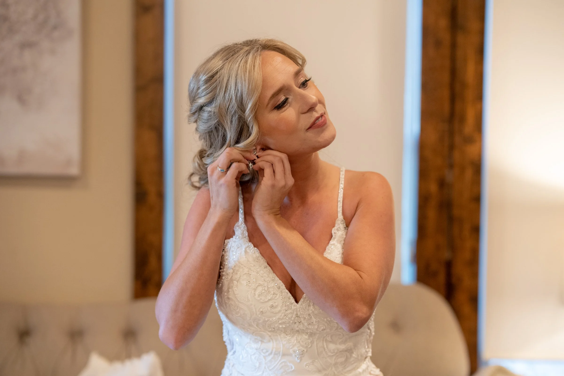 A woman with blonde hair, dressed in a white lace dress, is putting on earrings in a room with wooden beams.