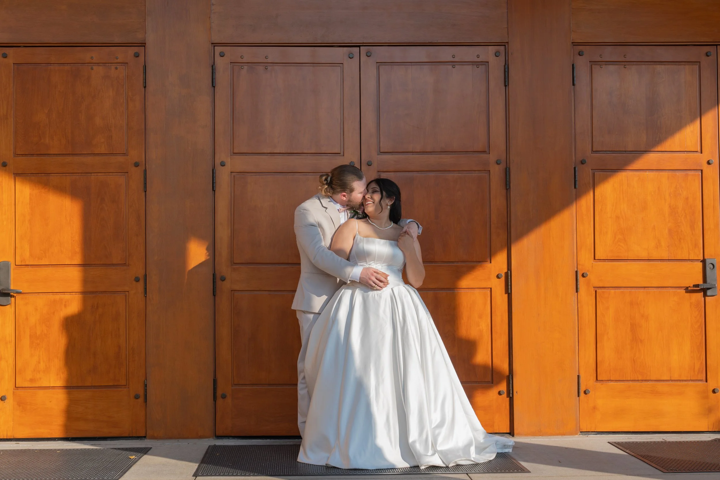 A bride and groom share a kiss in front of wooden doors. The groom wears a light-colored suit, and the bride wears a white wedding dress with a pearl necklace.