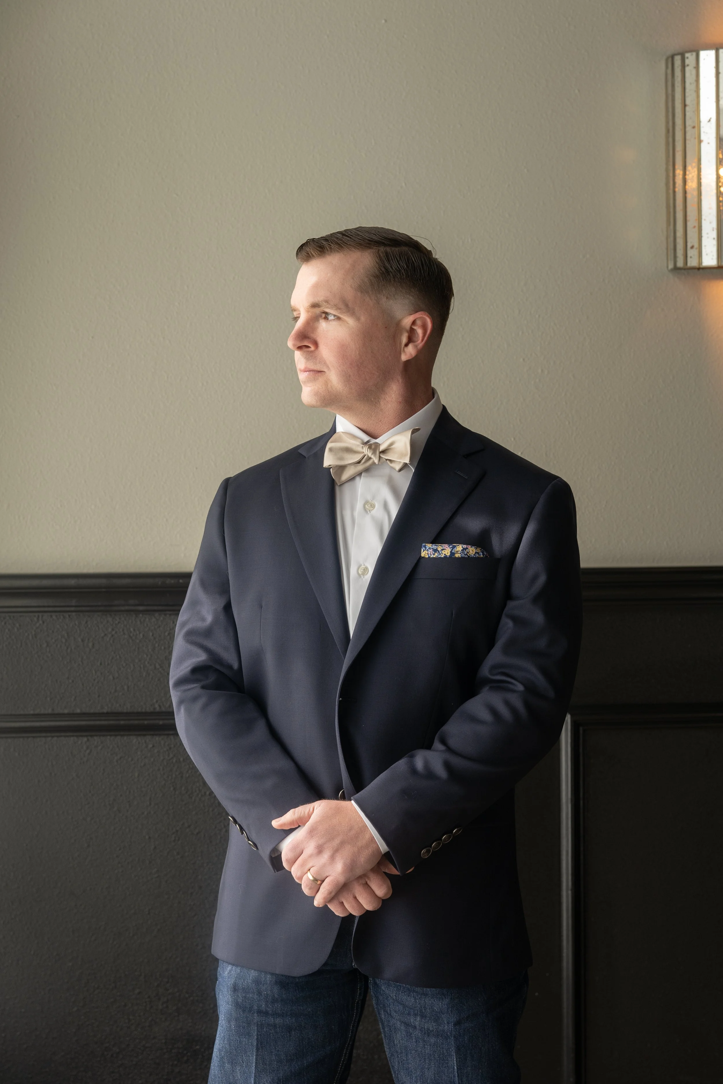 A man in a navy blue tuxedo with a cream bowtie and pocket square, standing against a beige wall with black trim, looking to his left with his hands clasped in front of him.