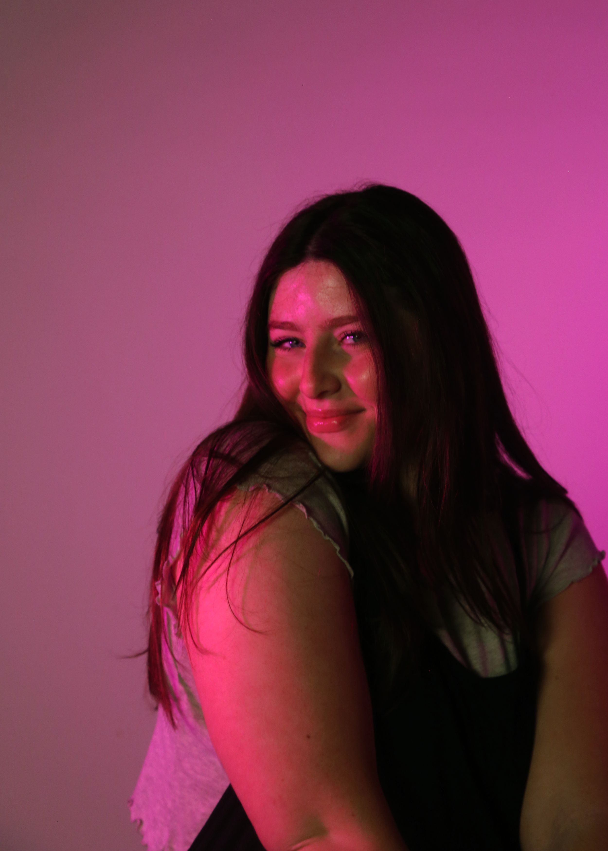 This portrait of Ava Shreffler continues the vibrant, personality-driven theme of the shoot. Using bold pink lighting to create dimension and mood, this image captures a more confident and playful expression. The soft pose and subtle smirk add attitu
