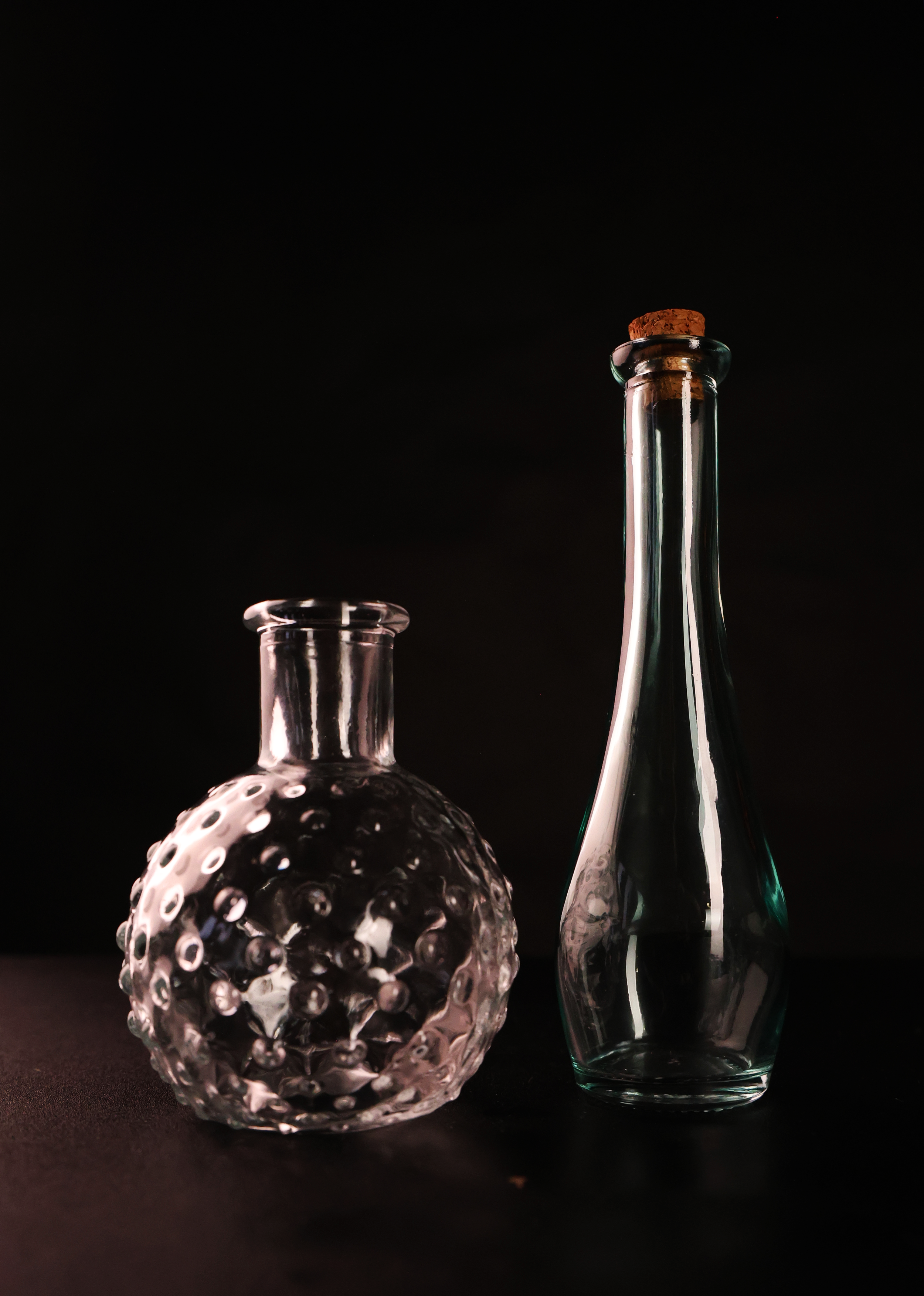 Featuring two transparent glass bottles photographed in a studio setting against a black background as part of a still life assignment. The goal of this setup was to explore shooting reflective, transparent glass in a darker environment without allow