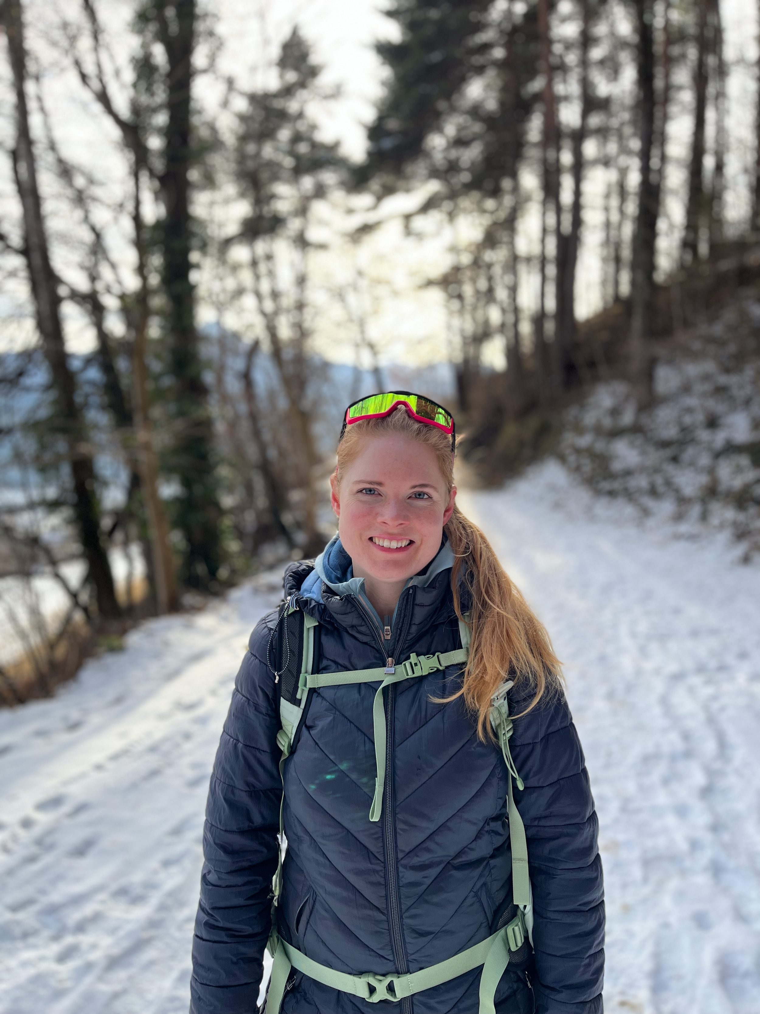 A smiling woman with long red hair, wearing sunglasses on her head, a navy blue jacket, and a backpack, standing on a snowy trail in a forest with trees and snow in the background.