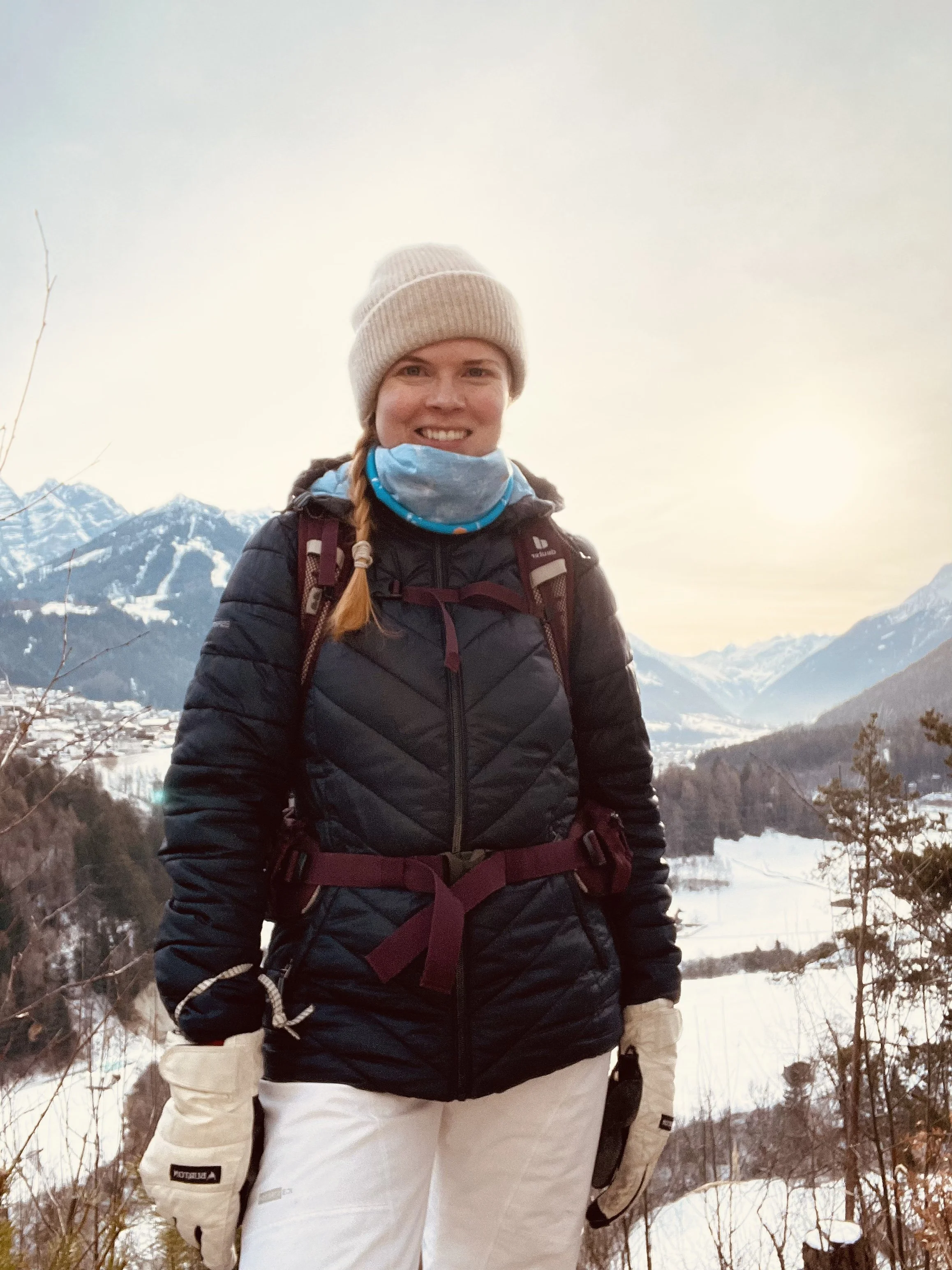A woman dressed in winter outdoor gear standing in a snowy mountain landscape with snow-covered peaks and trees in the background.