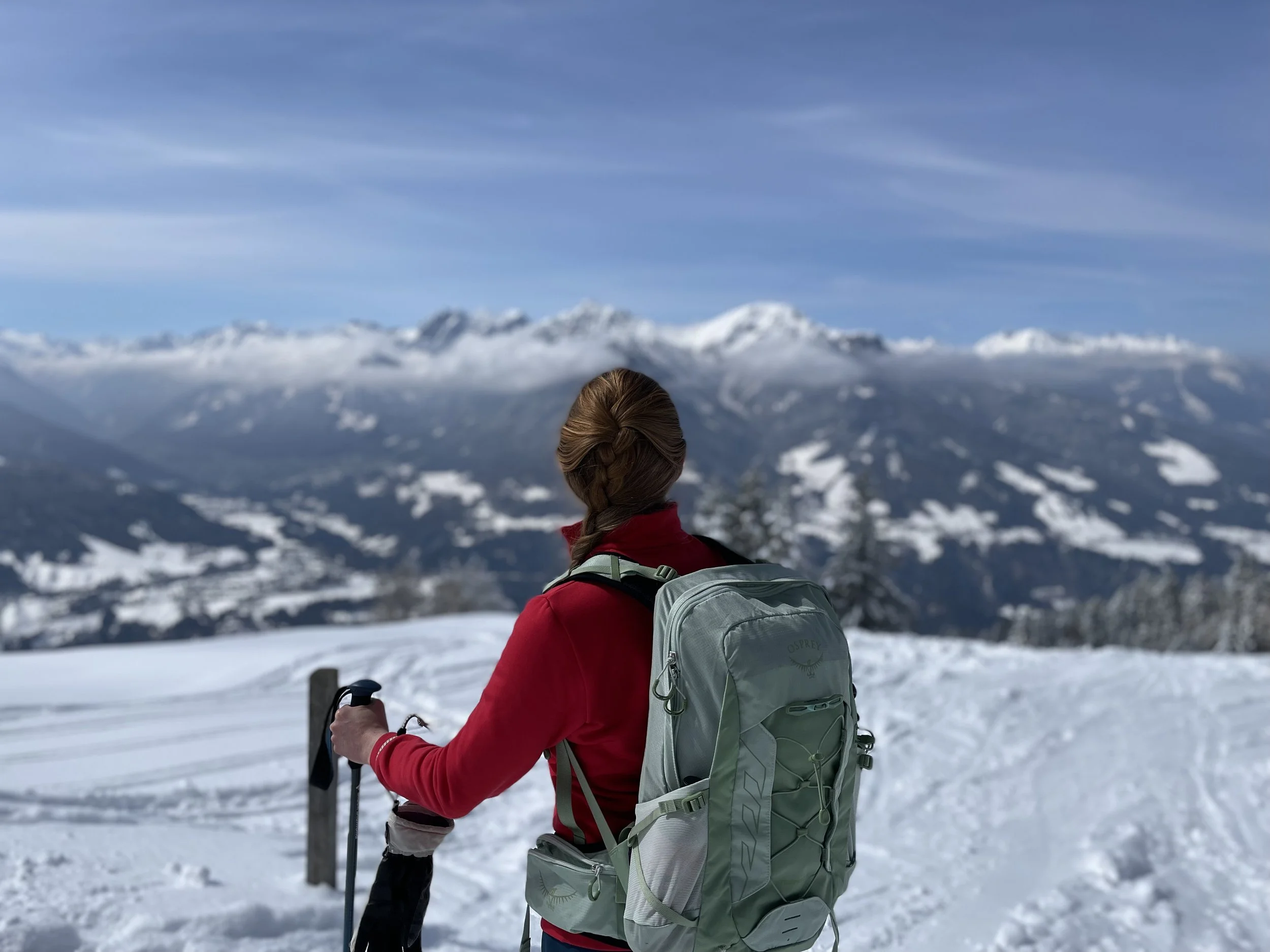 Woman with a braid in her red hair, wearing a red long-sleeved sweater and a green backpack. In the background, you can see snow-covered mountains.