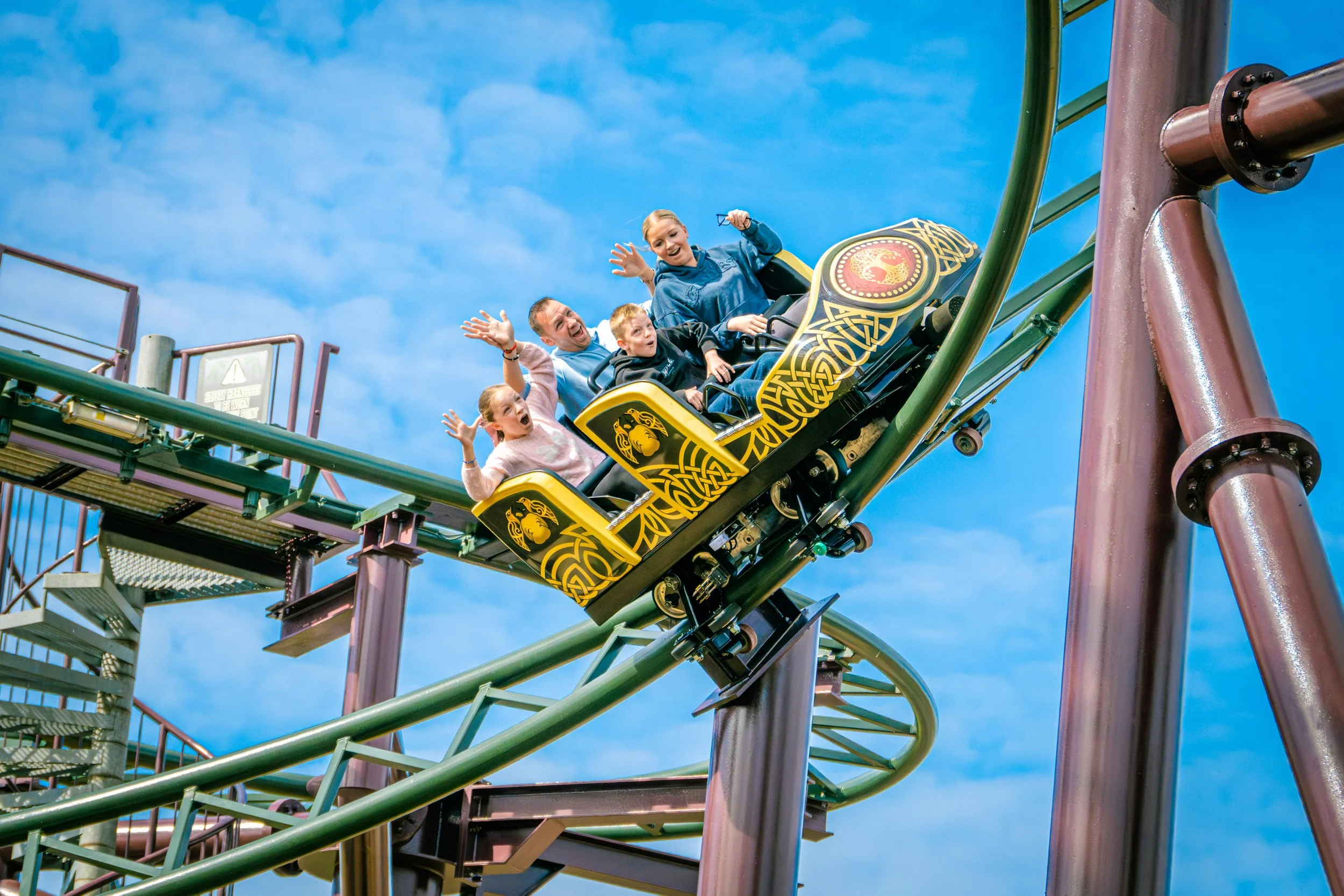 A group of five people riding a roller coaster at an amusement park. They appear excited and happy, with some waving and smiling. The roller coaster track is green, and the train has a yellow and black design with emblem. The sky is clear and blue, with some clouds visible.