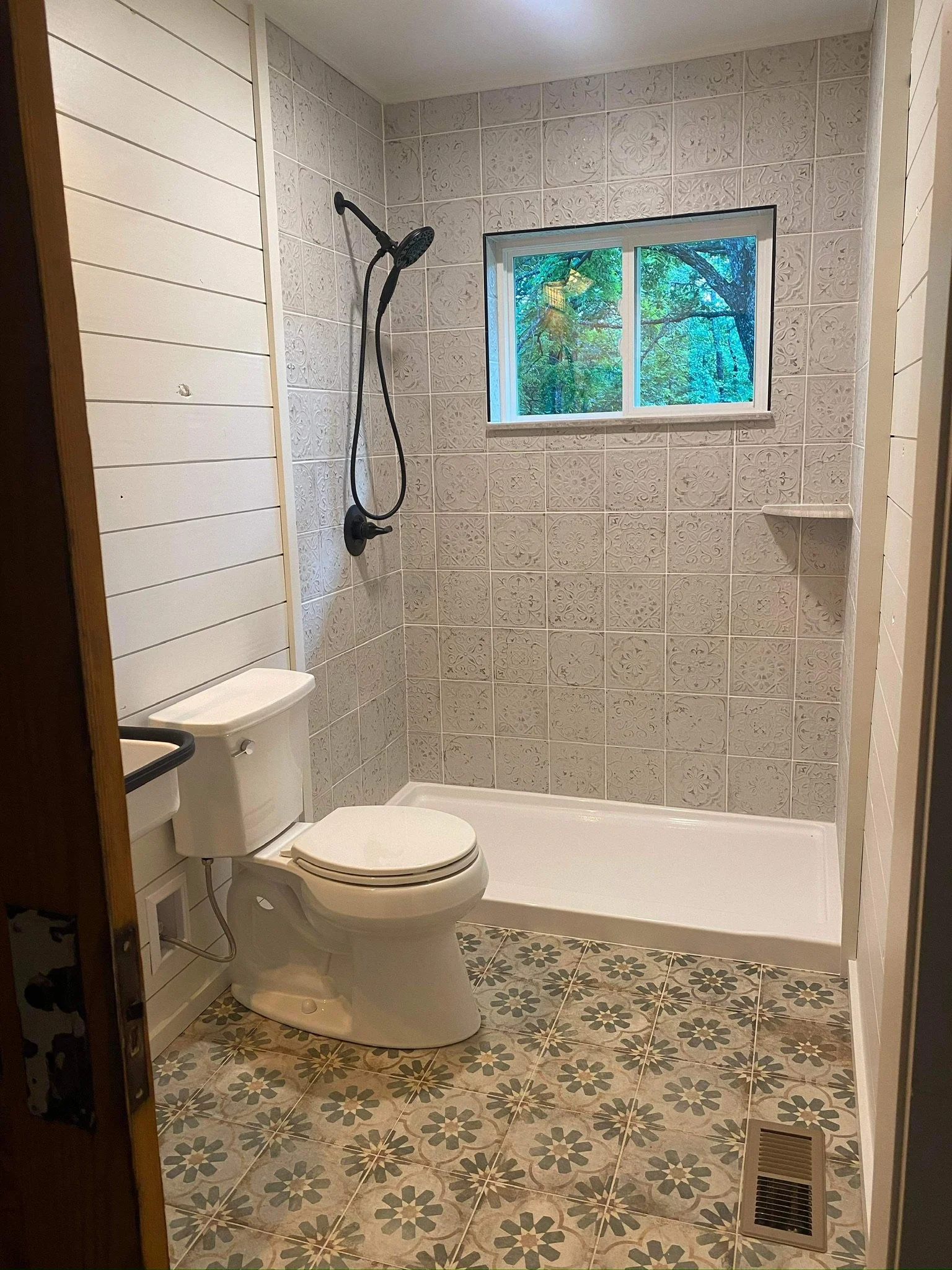Bathroom with patterned floor tiles, white toilet, shower area with black showerhead, small window showing greenery outside, and white wall paneling.