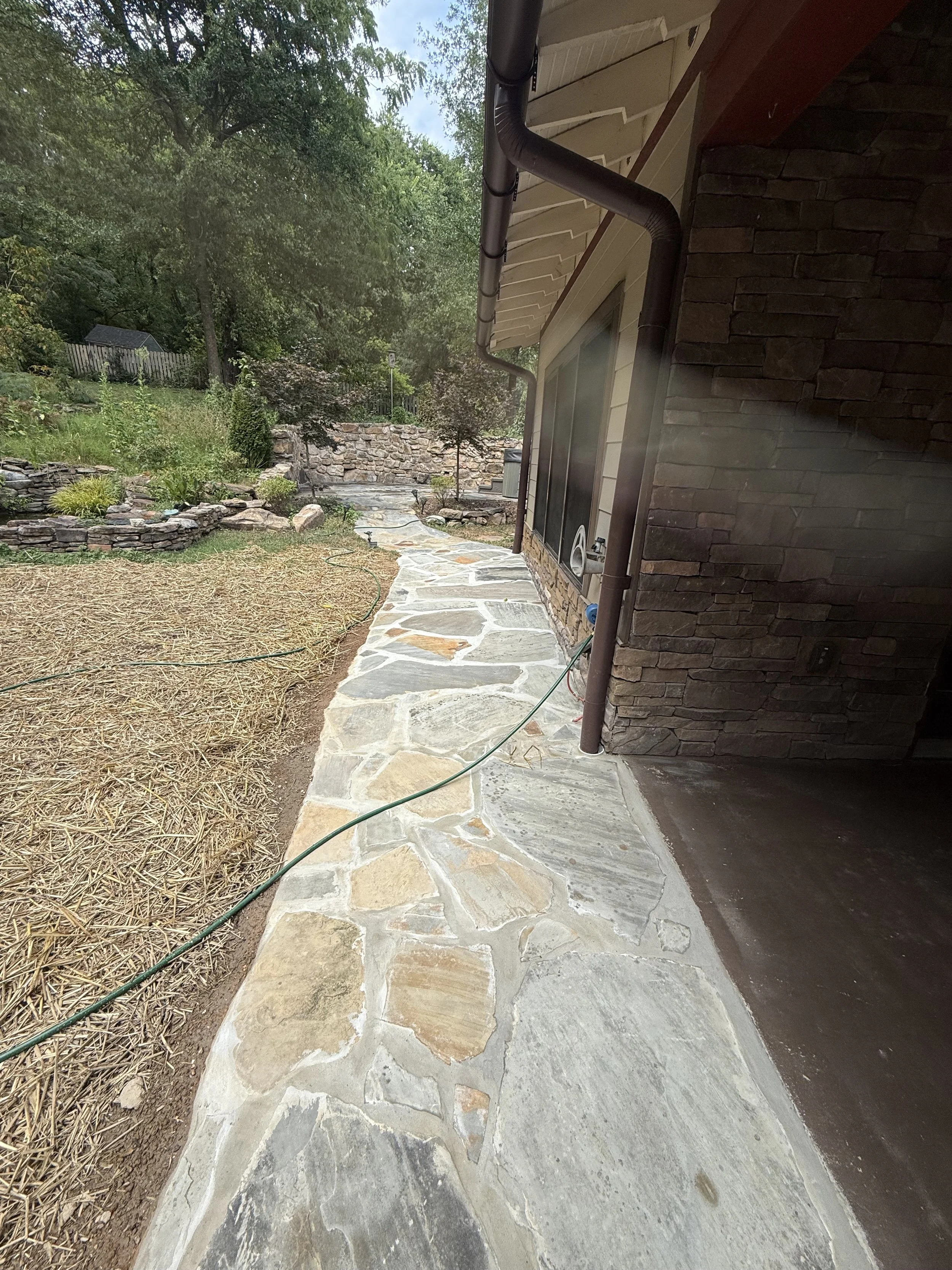 A stone pathway runs alongside a house with a brick exterior wall, and a brown gutter downspout. There is a garden area with plants and rocks on the left, and trees in the background.