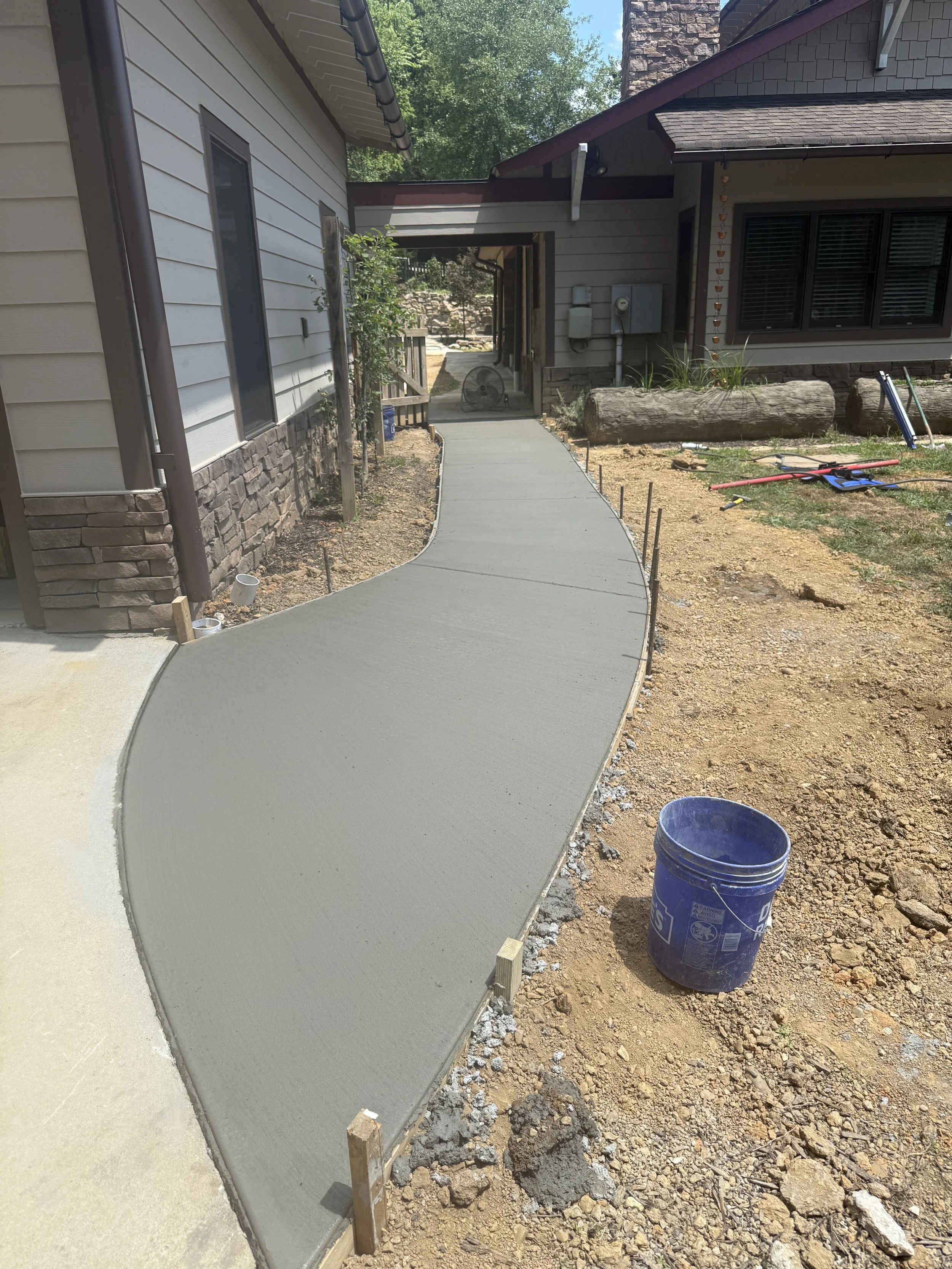 Freshly poured concrete walkway in front of a house, with wooden form boards and reinforcements, under construction on a sunny day.