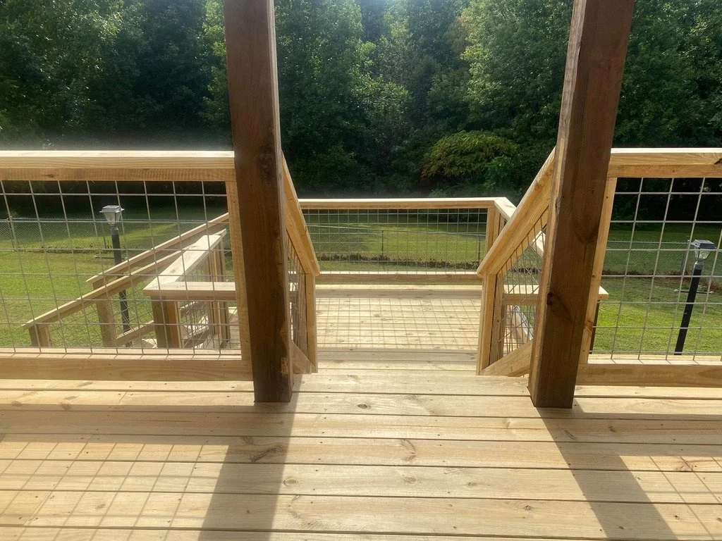 Looking down a wooden outdoor staircase with metal wire railings, leading to a grassy yard and trees beyond.