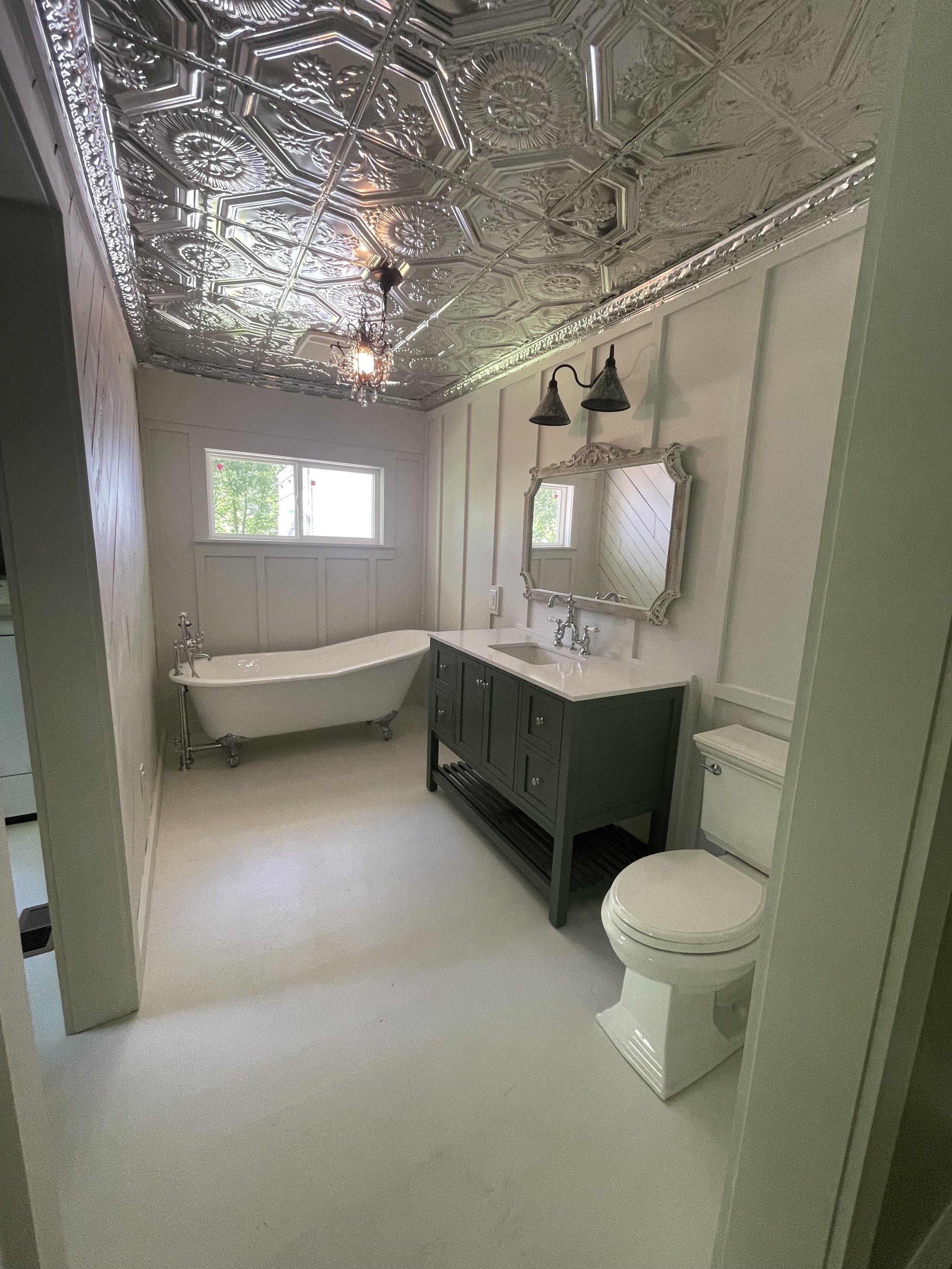 A vintage-style bathroom with a silver decorative tin ceiling, a white clawfoot bathtub, a dark green vanity with a mirror, a white toilet, and a window with outside greenery visible.