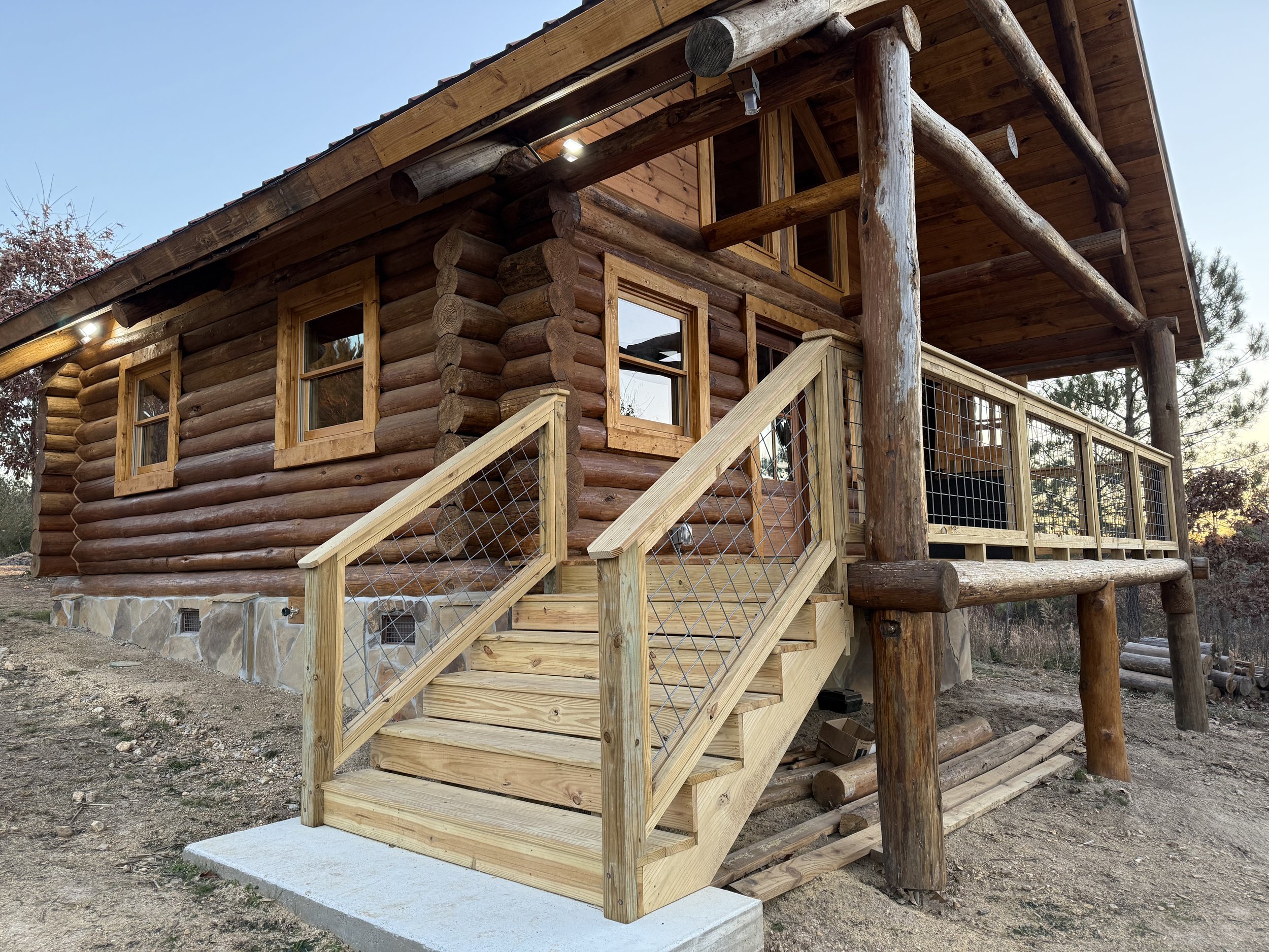 Log cabin under construction with new wooden stairs and railing leading to a porch, surrounded by a natural landscape during dusk.