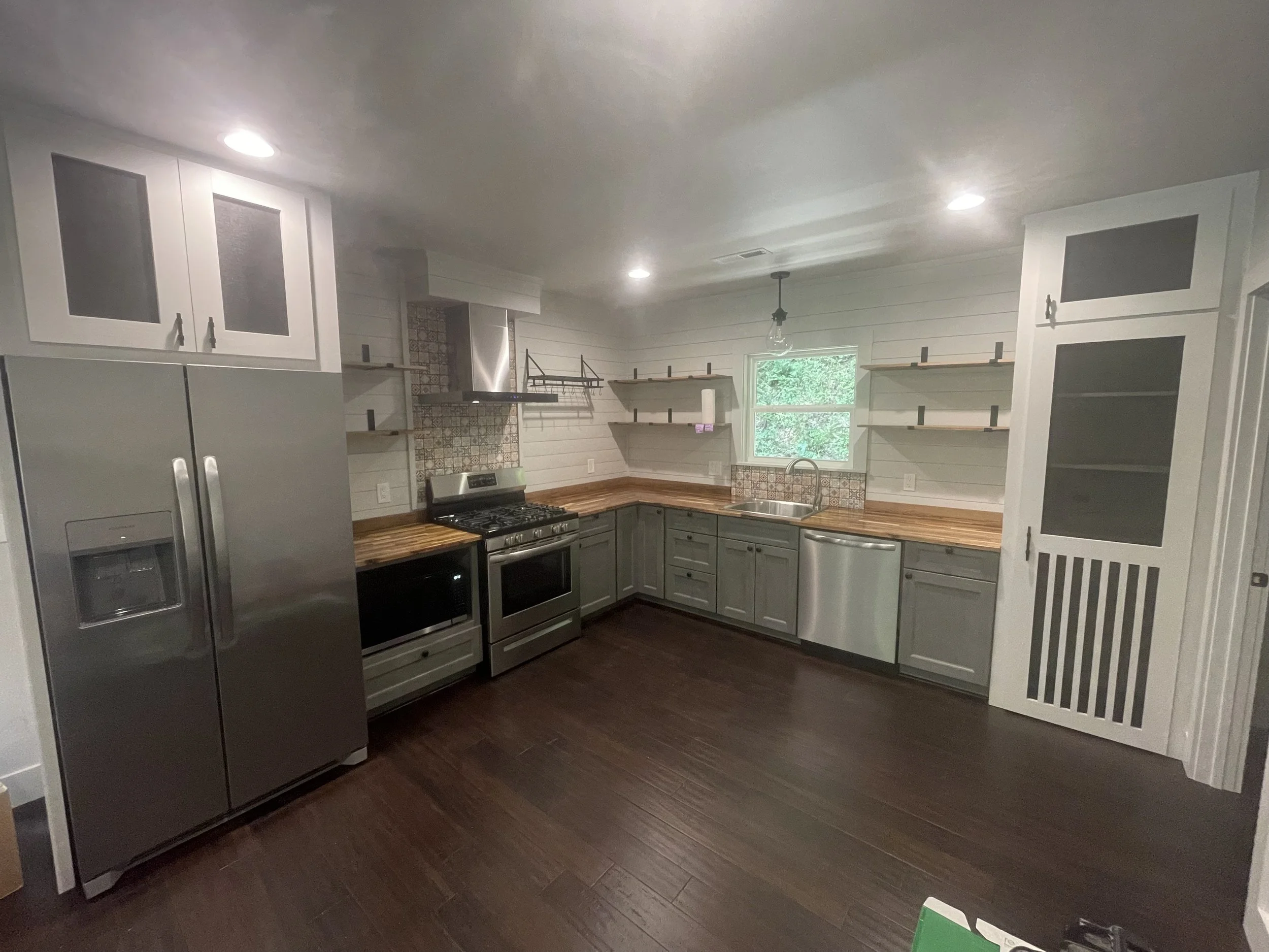 Modern kitchen with gray cabinets, stainless steel appliances, open wooden shelves, and a small window showing greenery outside.