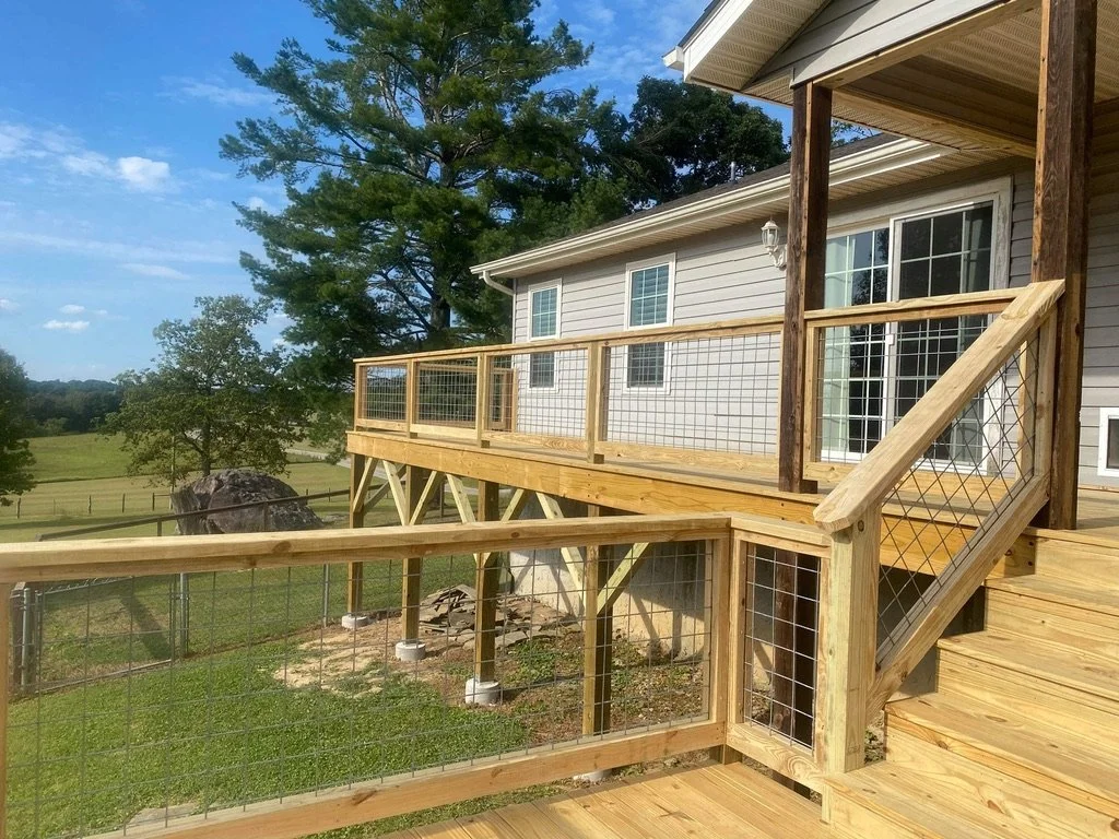 Backyard deck with wooden railings and fencing, attached to a house with sliding glass door, overlooking a grassy yard with trees and large rocks.