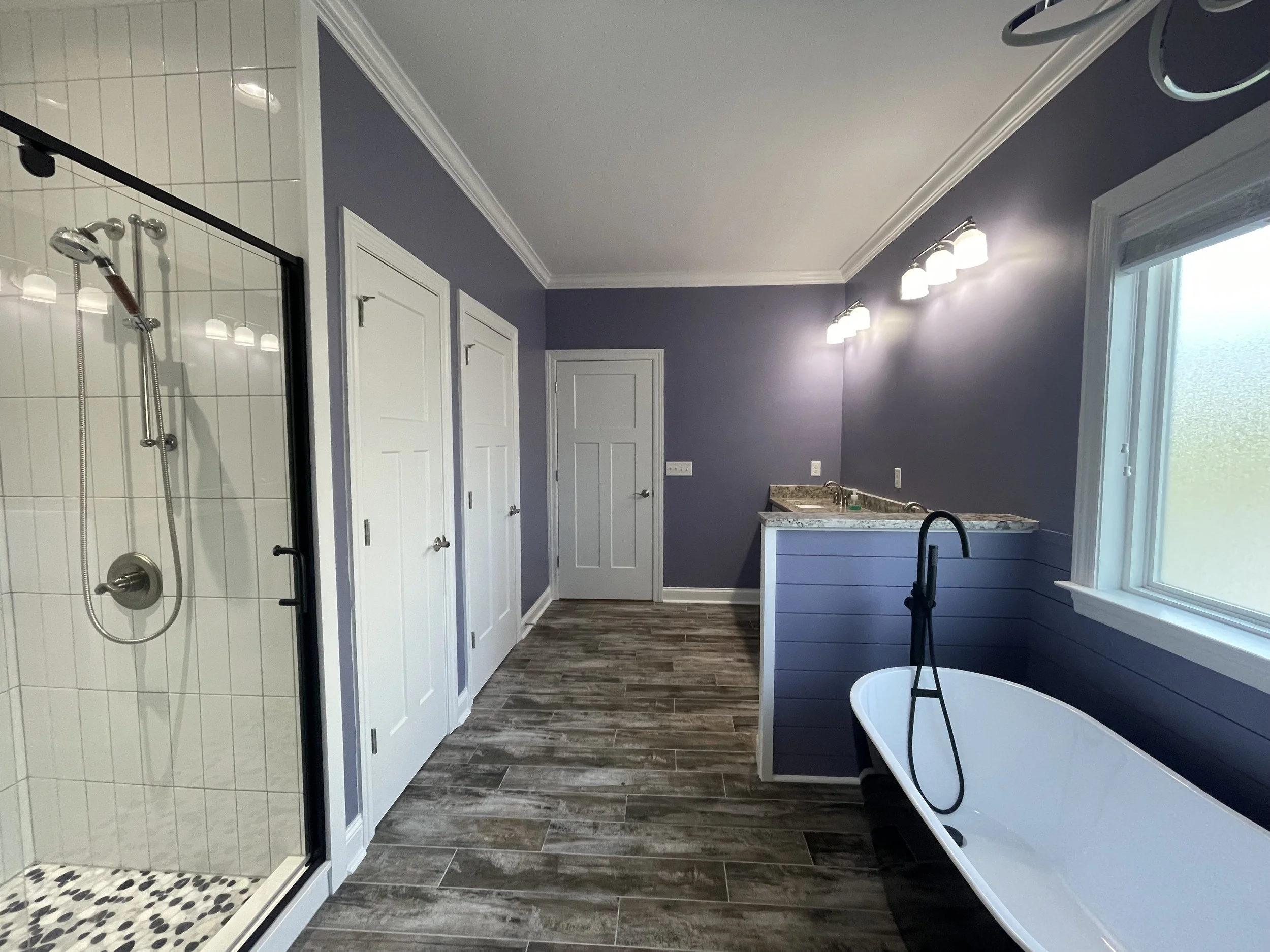 Bathroom with a walk-in shower, multiple white doors, a purple accent wall, a window with frosted glass, a black clawfoot bathtub with a garden hose, and a marble countertop with lighting fixtures.