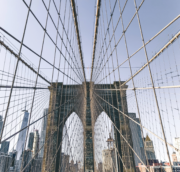 View of the Brooklyn Bridge from below, showing its stone towers and web of suspension cables against a clear sky.