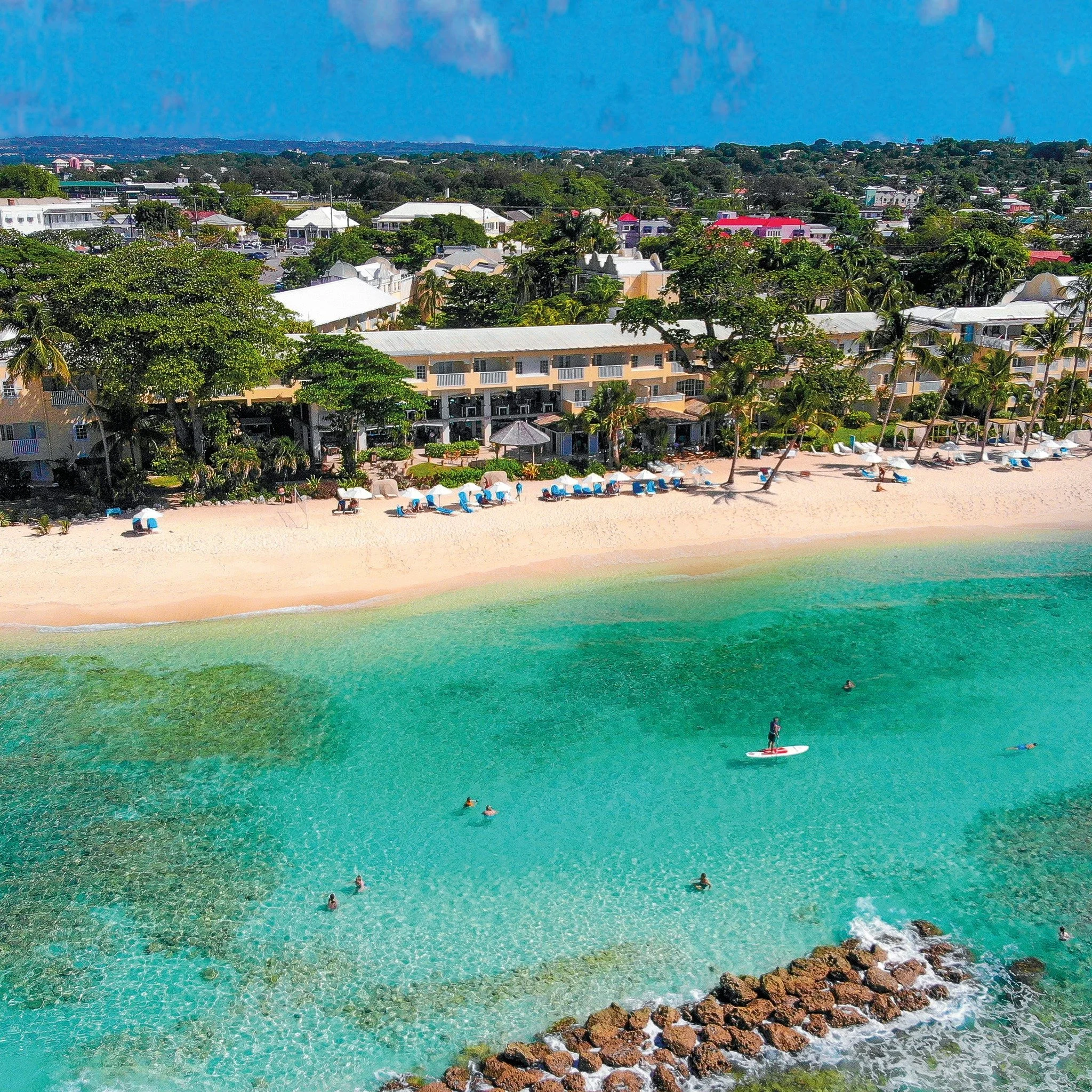 Beach with clear turquoise water, white sandy shore, and people swimming and surfing. There are lounge chairs with umbrellas along the beach and a rocky section near the water. In the background, there are trees and buildings.
