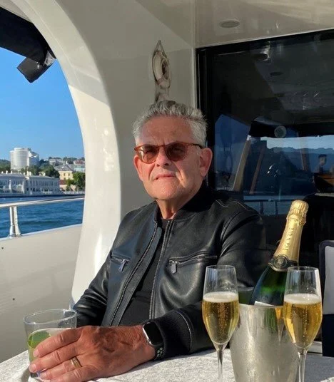 An older man wearing sunglasses and a black leather jacket sitting at a table on a boat with a bottle of champagne and two glasses of champagne, with water and buildings visible in the background.