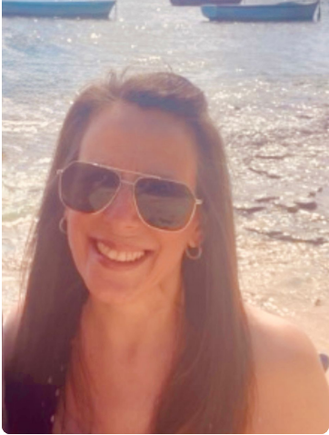 A woman with long brown hair wearing sunglasses, smiling, at the beach with water and boats in the background.
