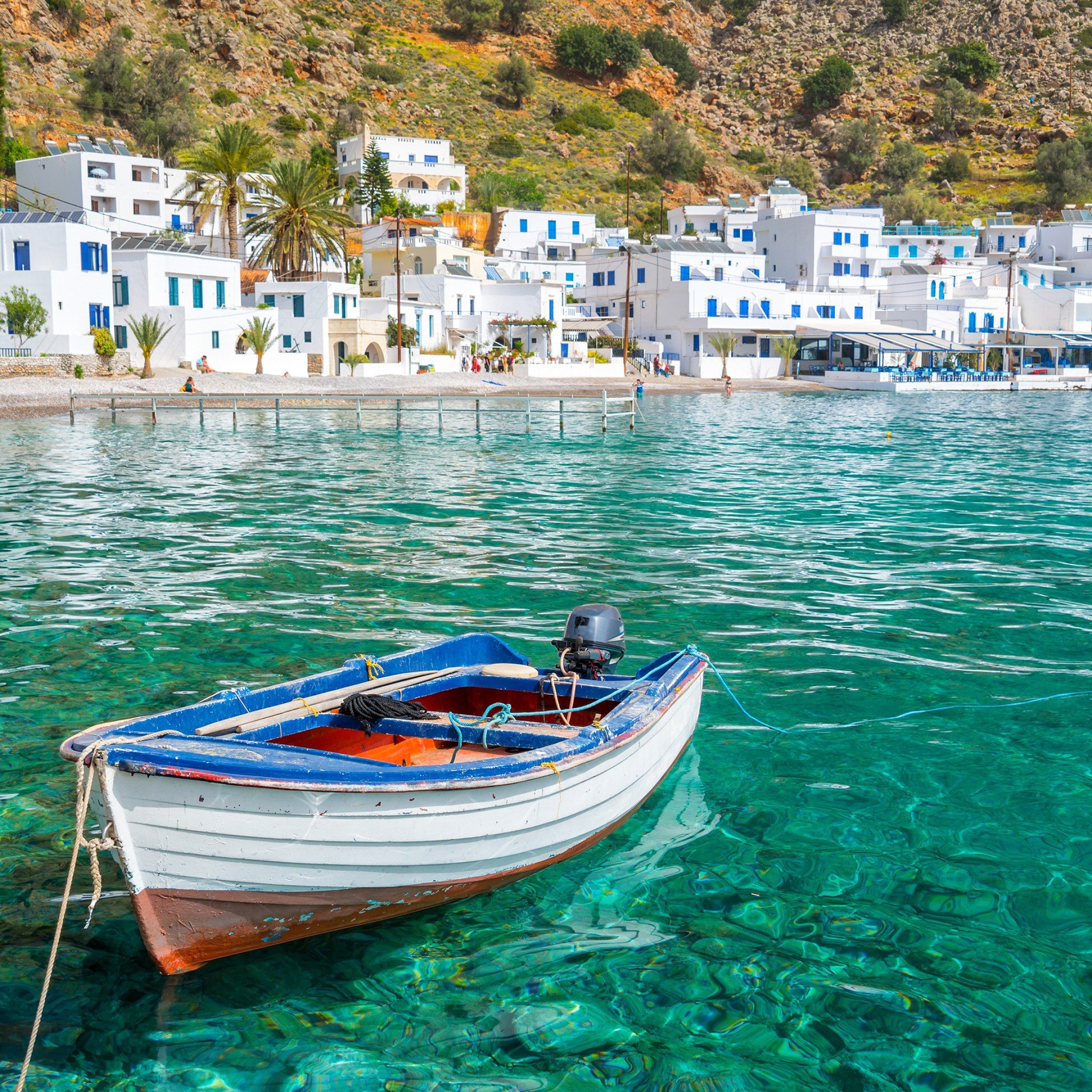 A small boat with a blue rim floats on clear green water near a coastal town with white buildings and blue accents, backed by a rocky hillside with sparse vegetation and palm trees.