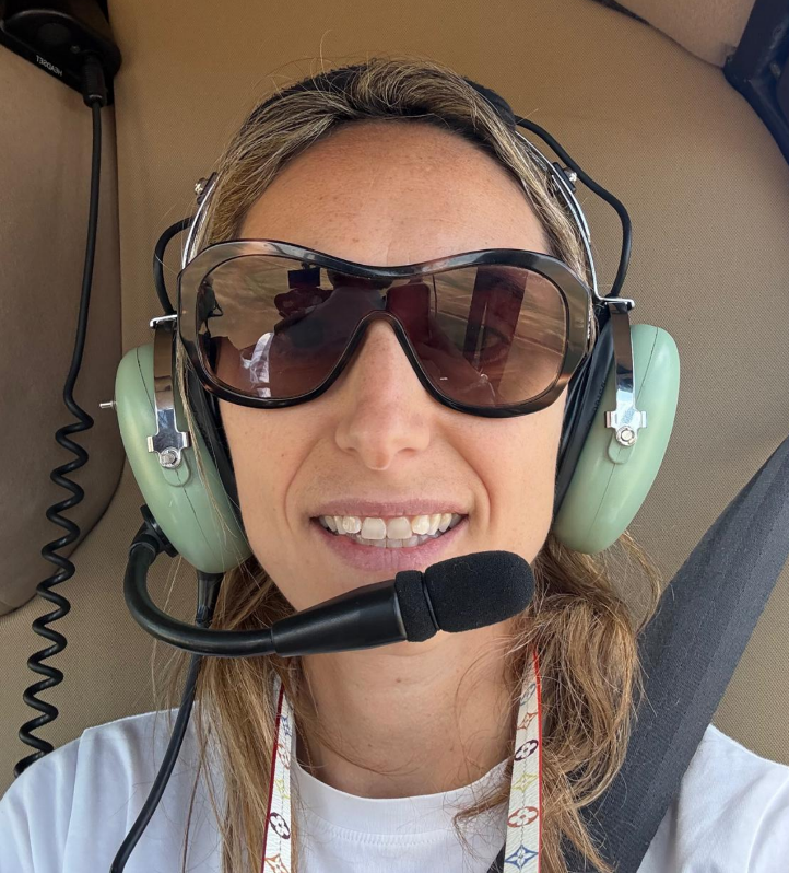 A woman wearing a headset with a microphone, large sunglasses, and a white shirt, sitting inside a helicopter cockpit.