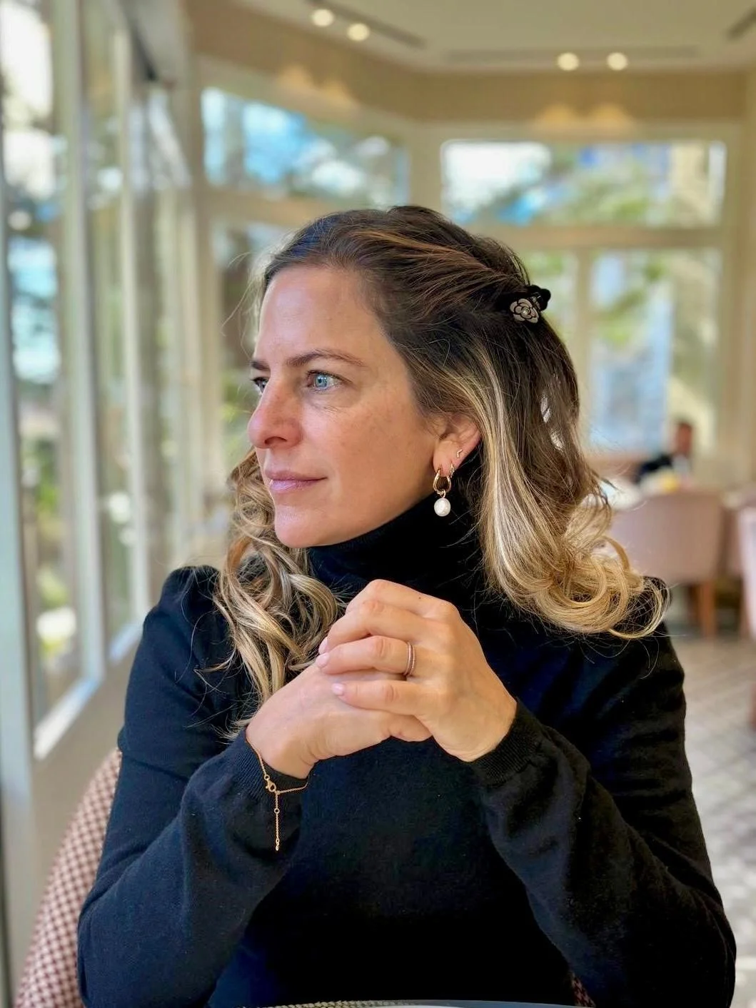 A woman with light brown curly hair, blue eyes, wearing a black turtleneck, pearl earrings, and a gold bracelet, sitting at a table in a well-lit restaurant with large windows and greenery outside.