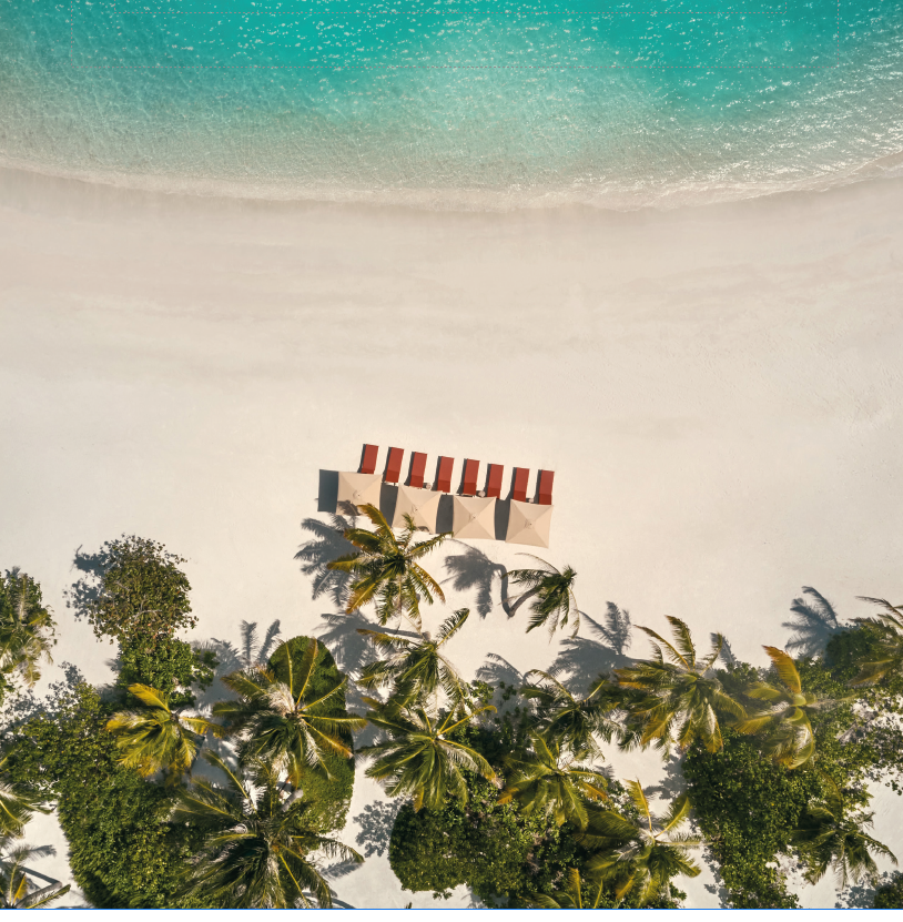 An aerial view of a tropical beach with a white sandy shoreline, turquoise water, and palm trees shading two beach umbrellas and lounge chairs.
