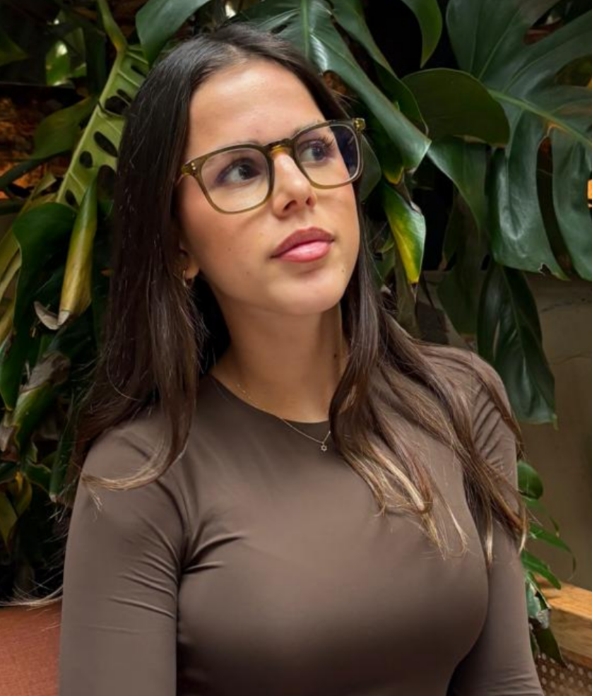 A woman with long dark hair and glasses looks thoughtfully to the side, sitting in front of large green tropical plants.