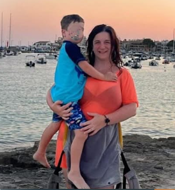 A woman holding a young boy on a rocky shoreline near the water at sunset, with boats and a marina in the background.