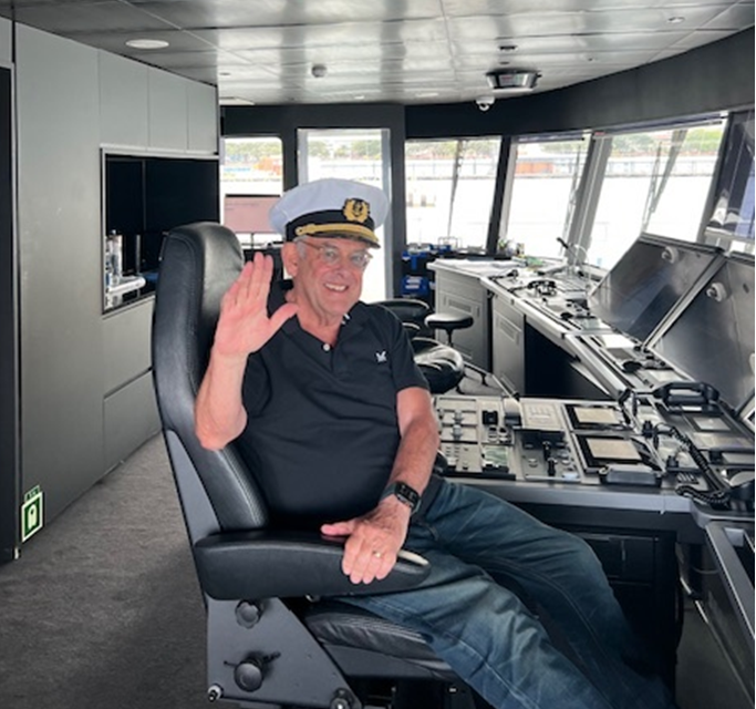 Older man wearing a captain's hat and black shirt, sitting in a ship's control room, smiling and waving.