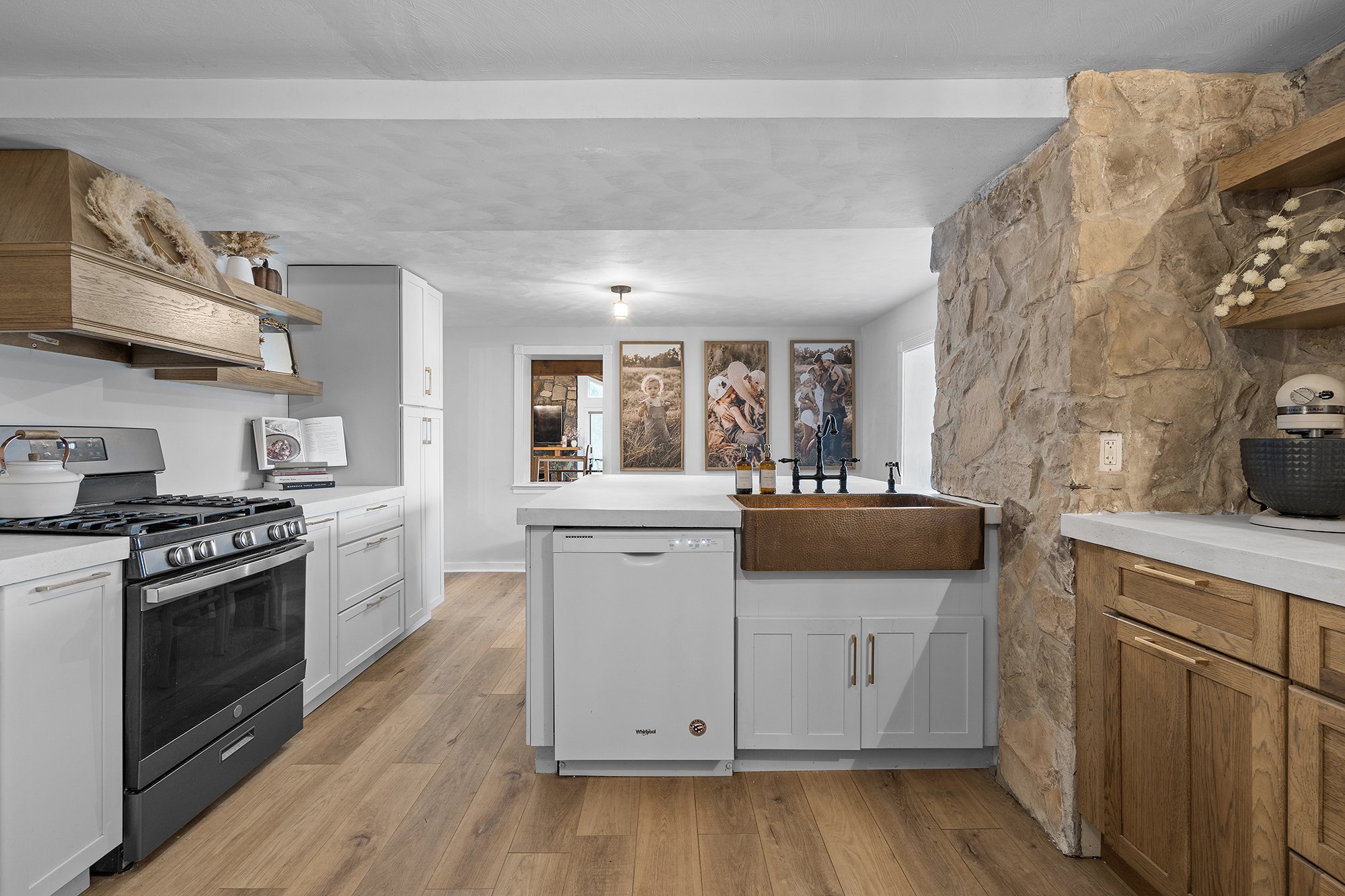 Modern kitchen with white cabinets, black stove, wooden shelves, a stone accent wall, and family photos on the wall in the background.