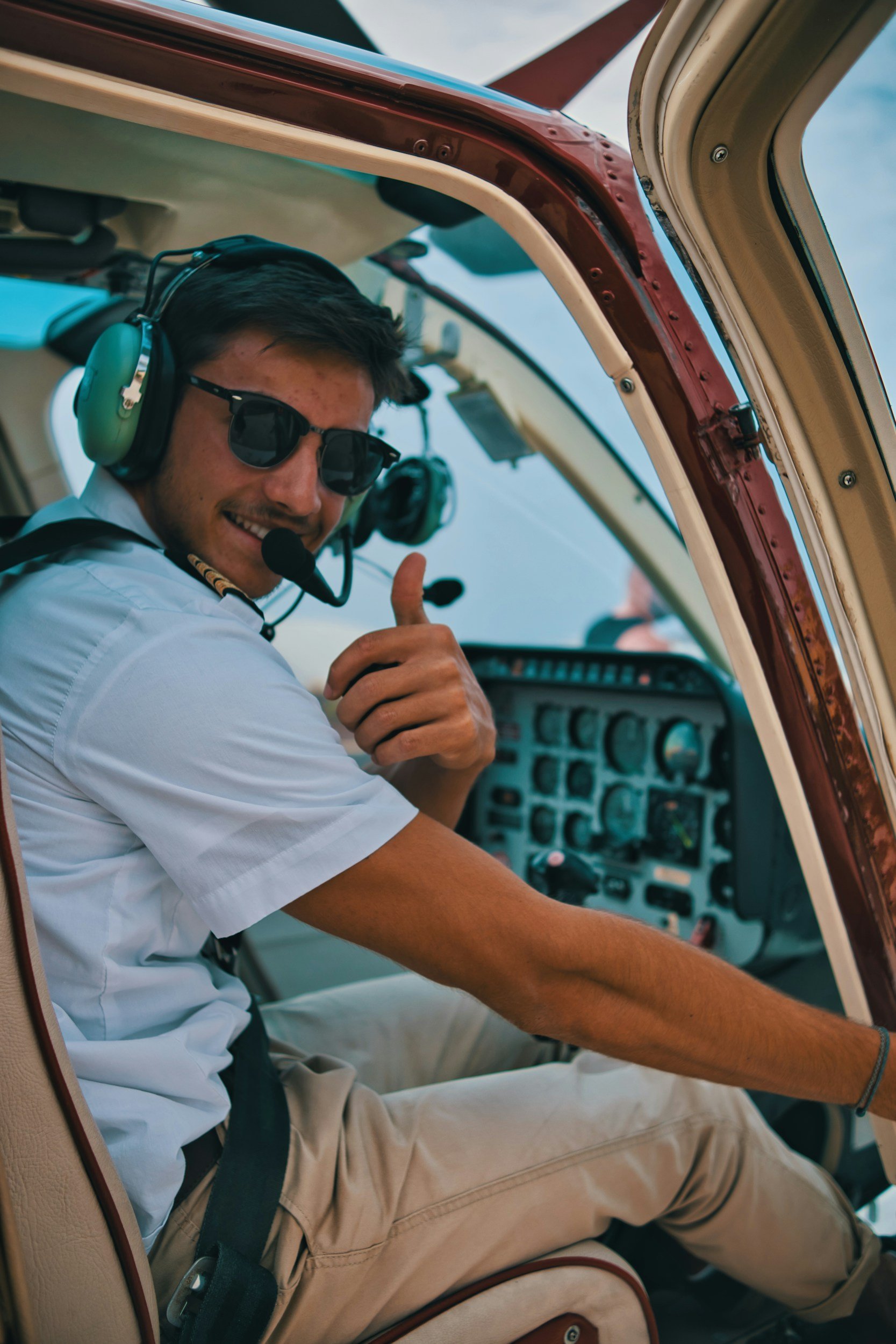 A smiling man wearing sunglasses and a headset, giving a thumbs-up while sitting in the cockpit of an airplane with visible controls and instruments.