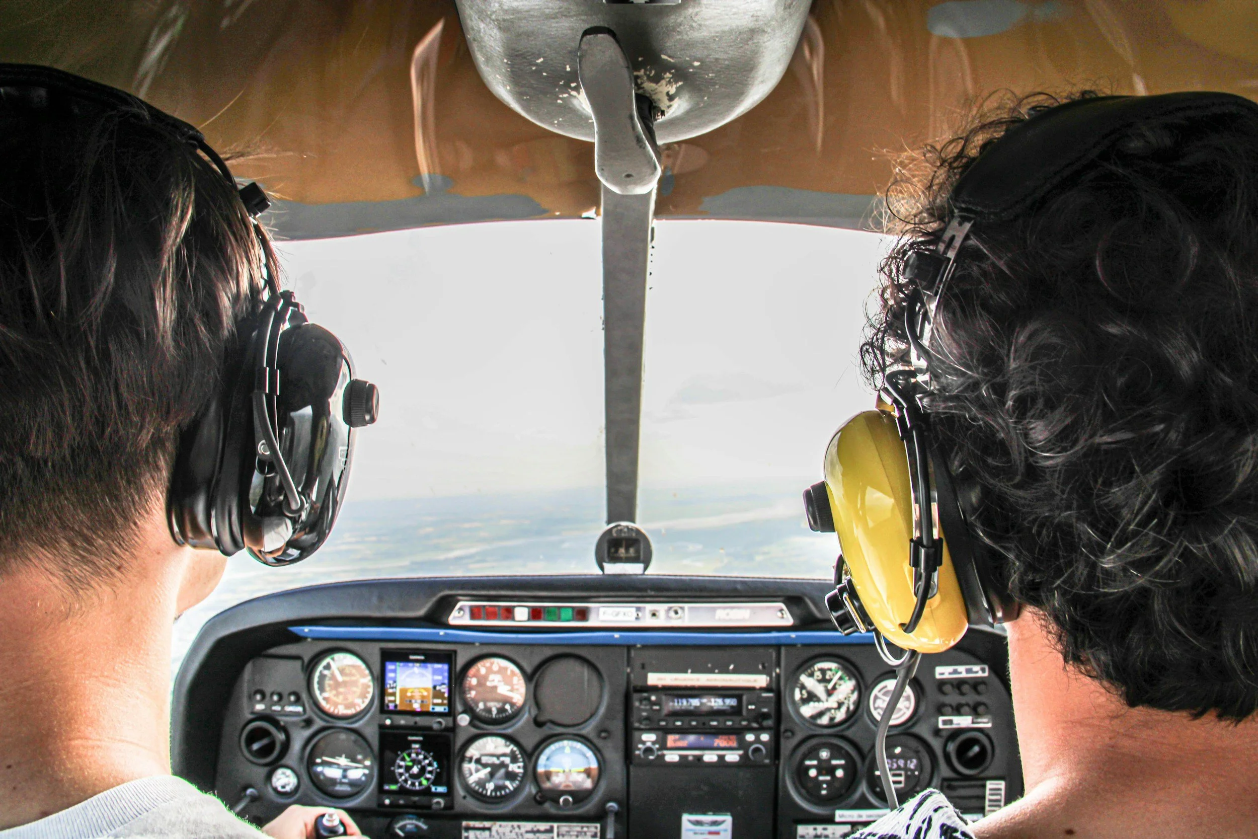The image shows the cockpit of a small aircraft with two people wearing headsets, flying over a flat landscape with clouds visible outside.