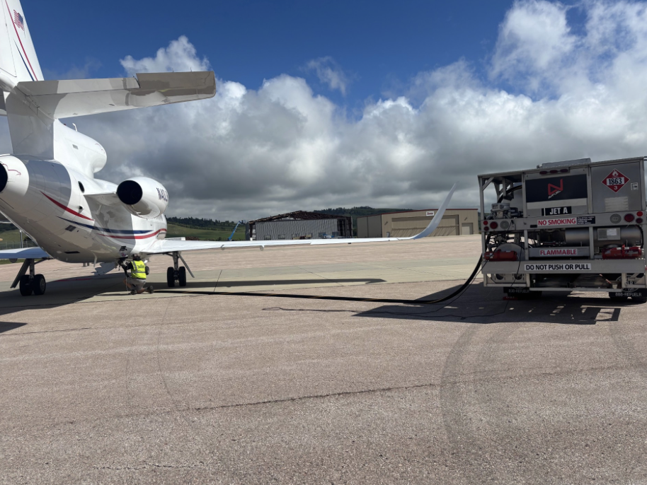 An airplane parked on the tarmac connected to a fuel truck with a hose, under partly cloudy skies.