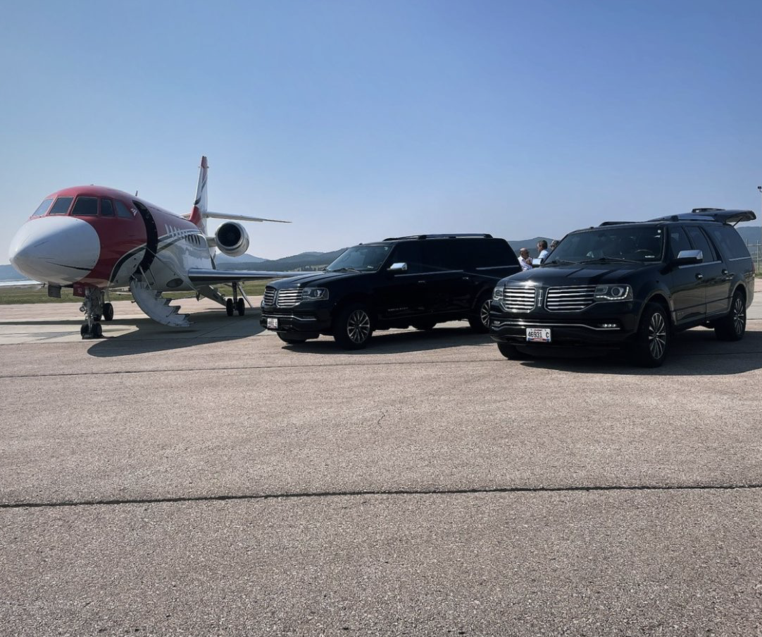 A private jet and two black SUVs parked on an airport tarmac on a clear day.