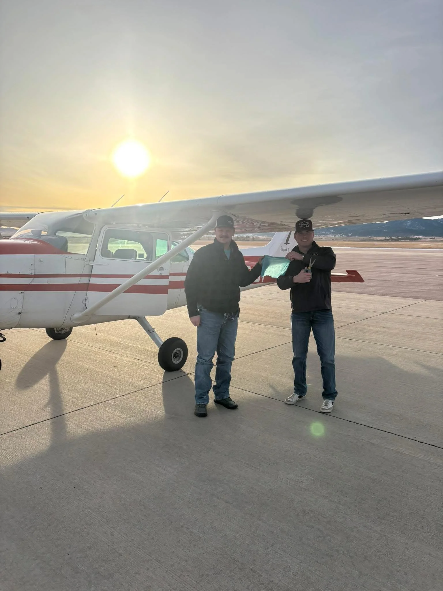 Two men standing in front of a small airplane on an airstrip during sunset, smiling, with clouds and mountains in the background.