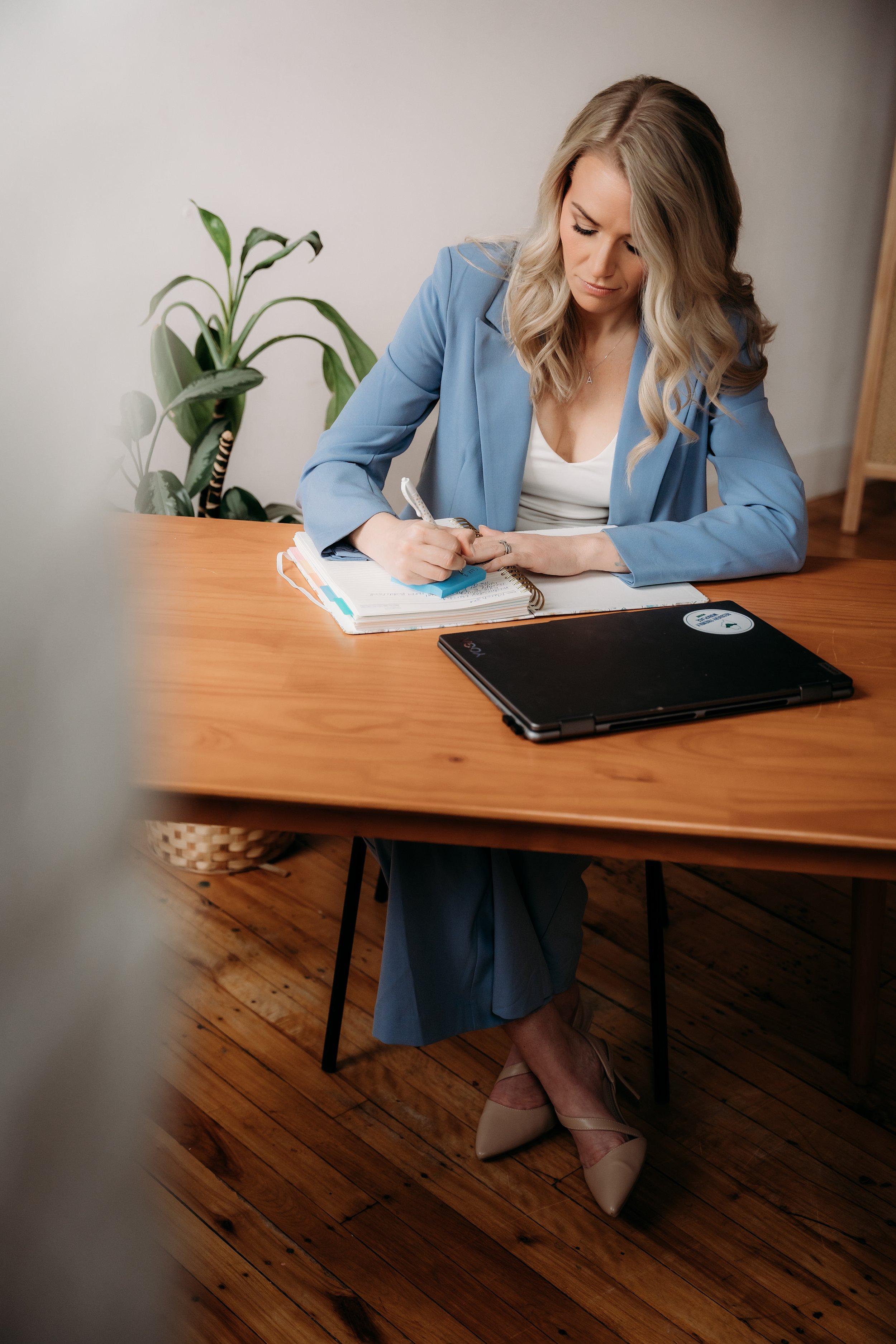 A woman in a light blue blazer and matching skirt is sitting at a wooden table, writing in a notebook with a pen. She has long, wavy blonde hair and is wearing beige high-heeled shoes. There is a black laptop on the table and a potted plant in the background.