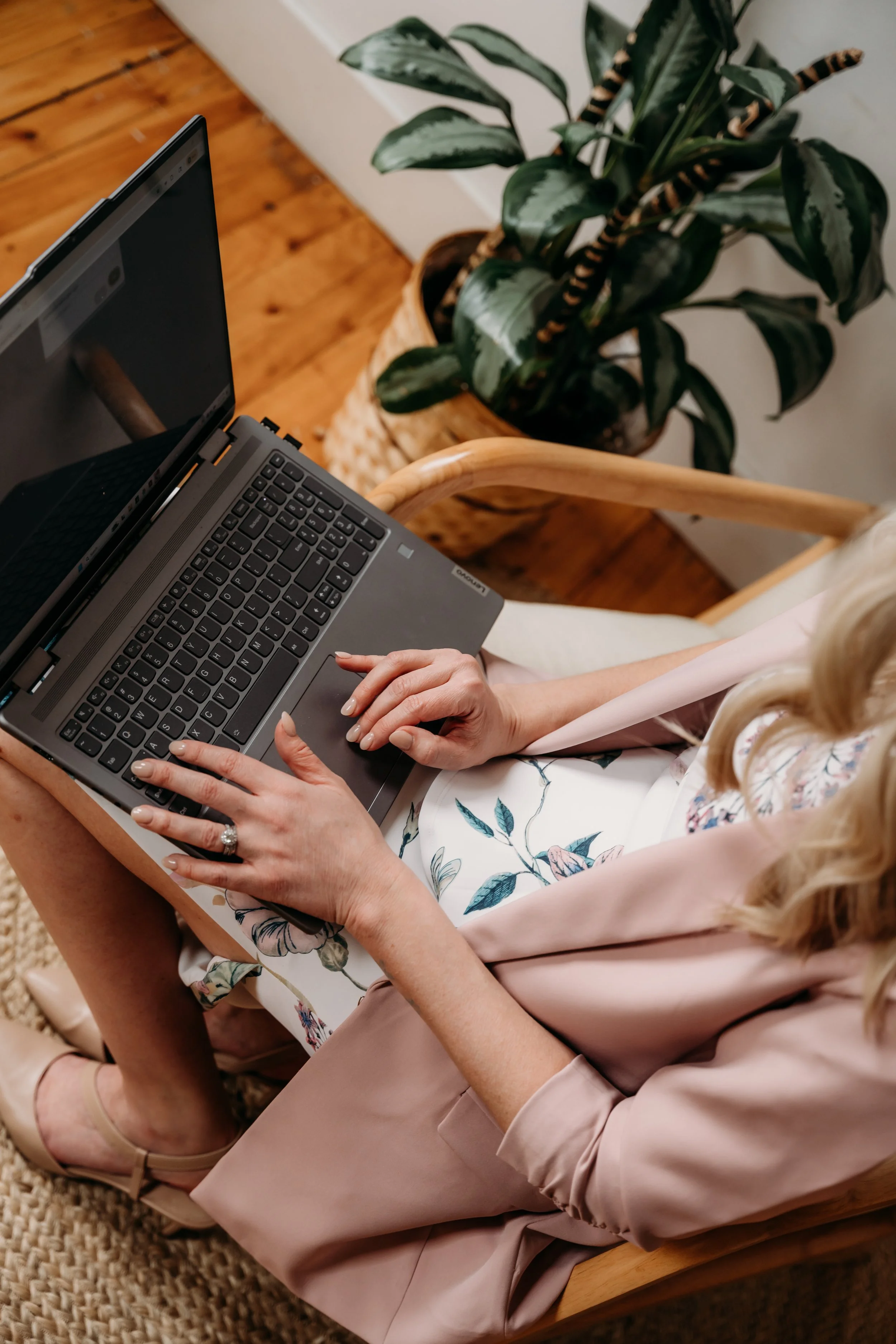 A woman sitting on a wooden chair using a laptop on her lap, wearing a pink floral dress, with a potted plant in the background.