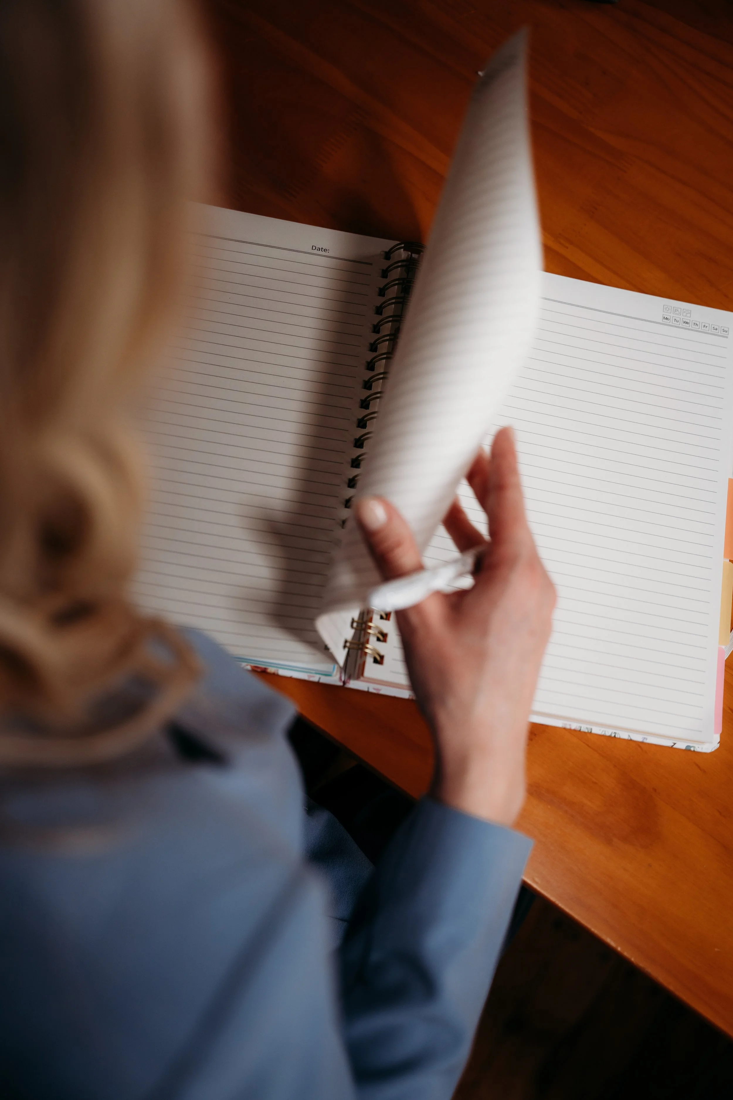 A woman with blonde hair in a ponytail is flipping through a spiral notebook with lined pages at a wooden table.