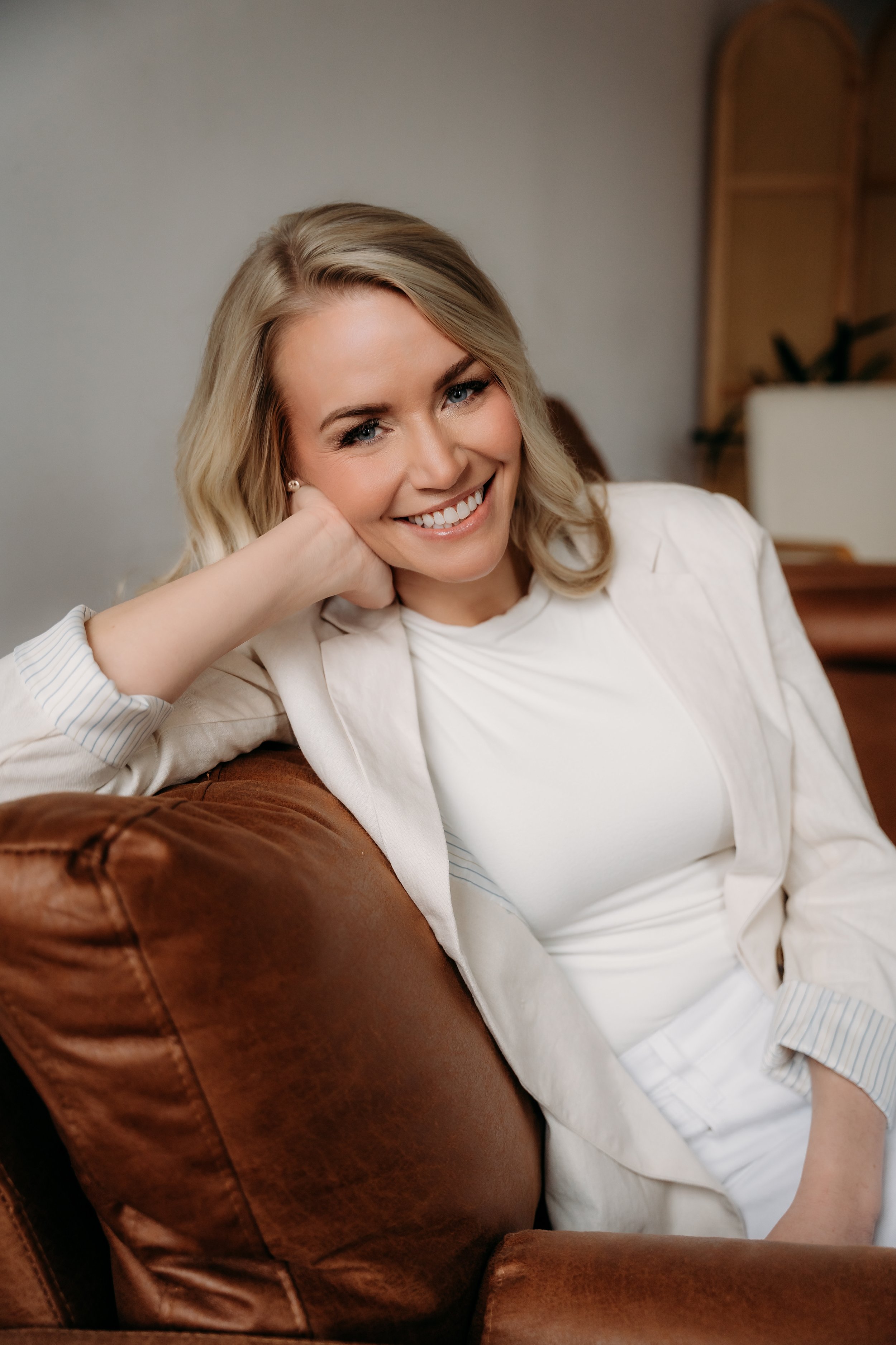 A smiling woman with blonde hair, wearing a white blazer and white top, leaning on her hand while sitting on a brown leather couch in a cozy room.
