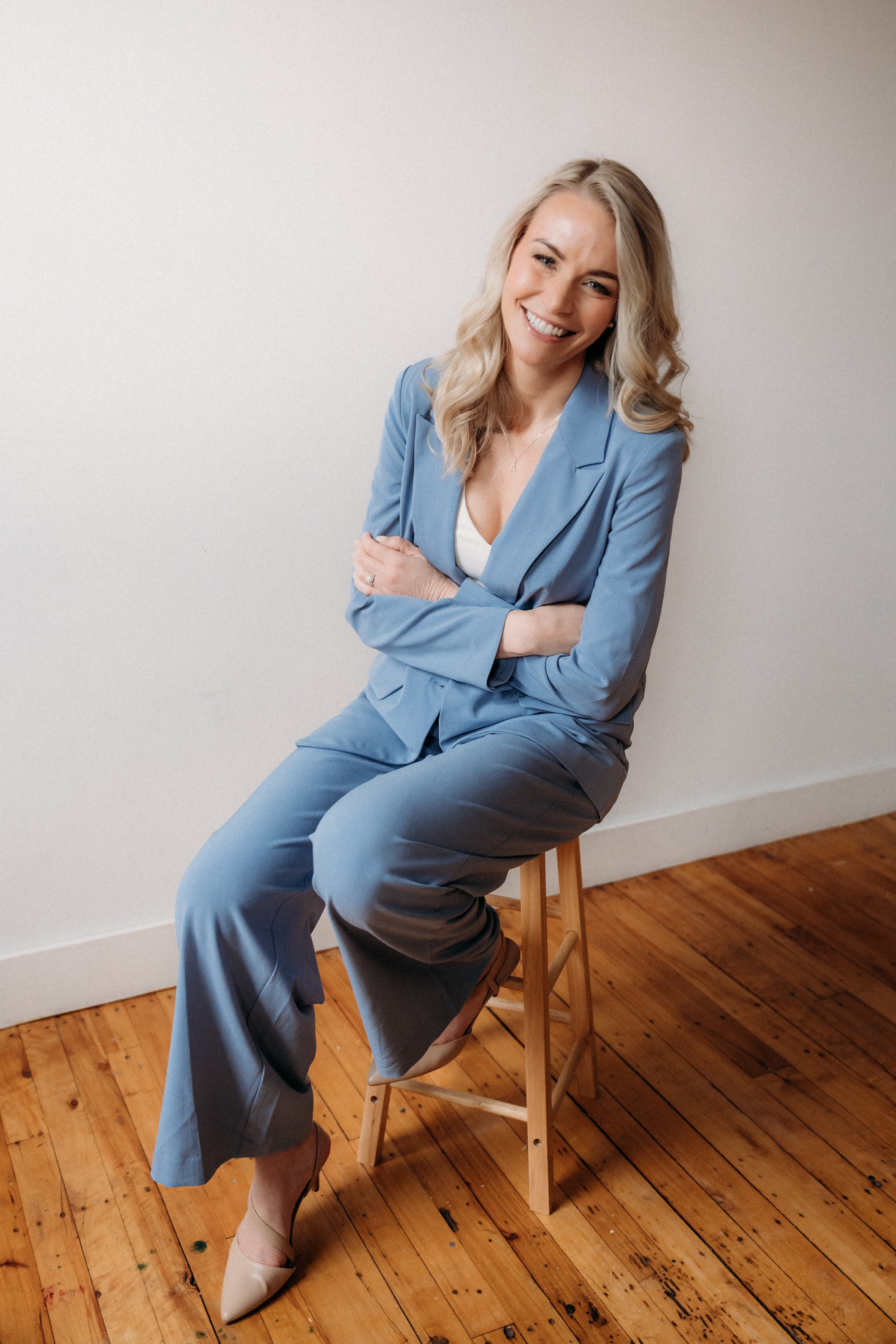 A smiling woman with blonde wavy hair sitting sideways on a wooden stool against a plain white wall. She is wearing a light blue blazer and matching pants, with a white top underneath and beige heels. Her arms are crossed, and her head is slightly tilted.