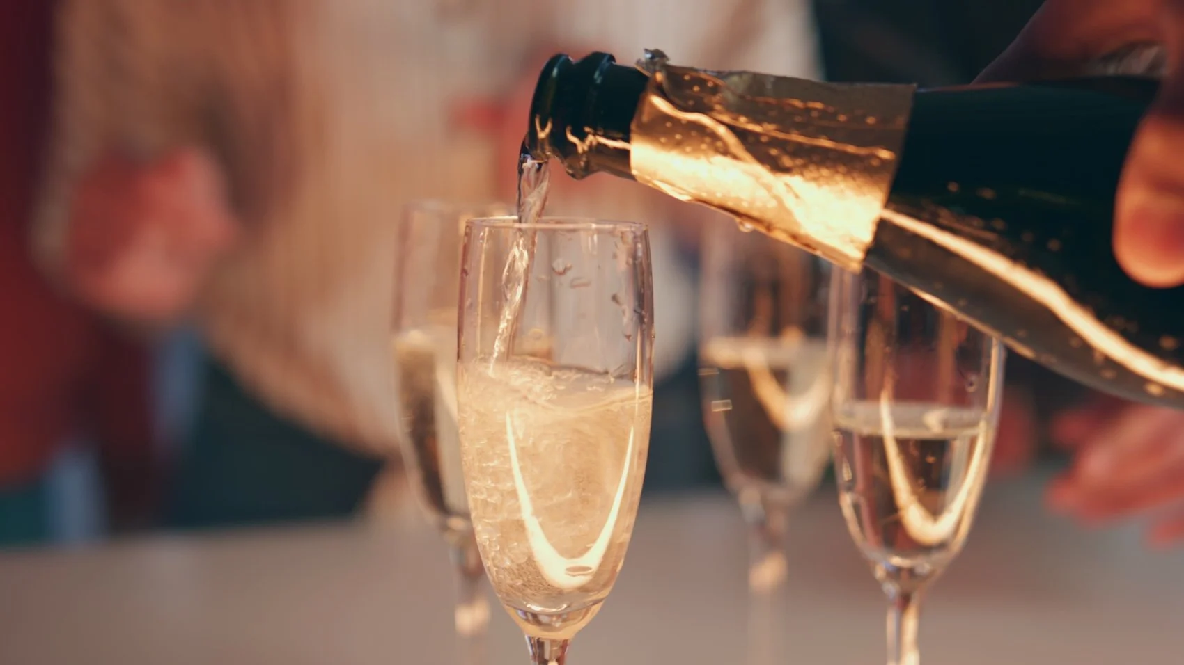 A close up of a bottle of champagne being poured into a class with a party in the background