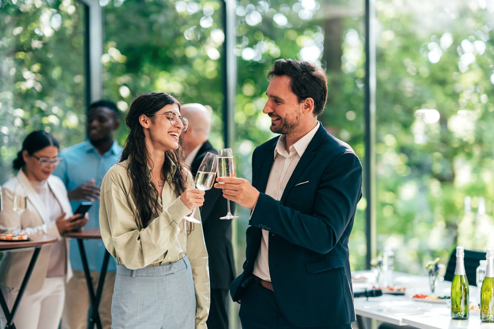 A man and a woman dressed in business casual clinking champagne glasses together in a corporate setting with big windows and a lot of light and greenery in the background