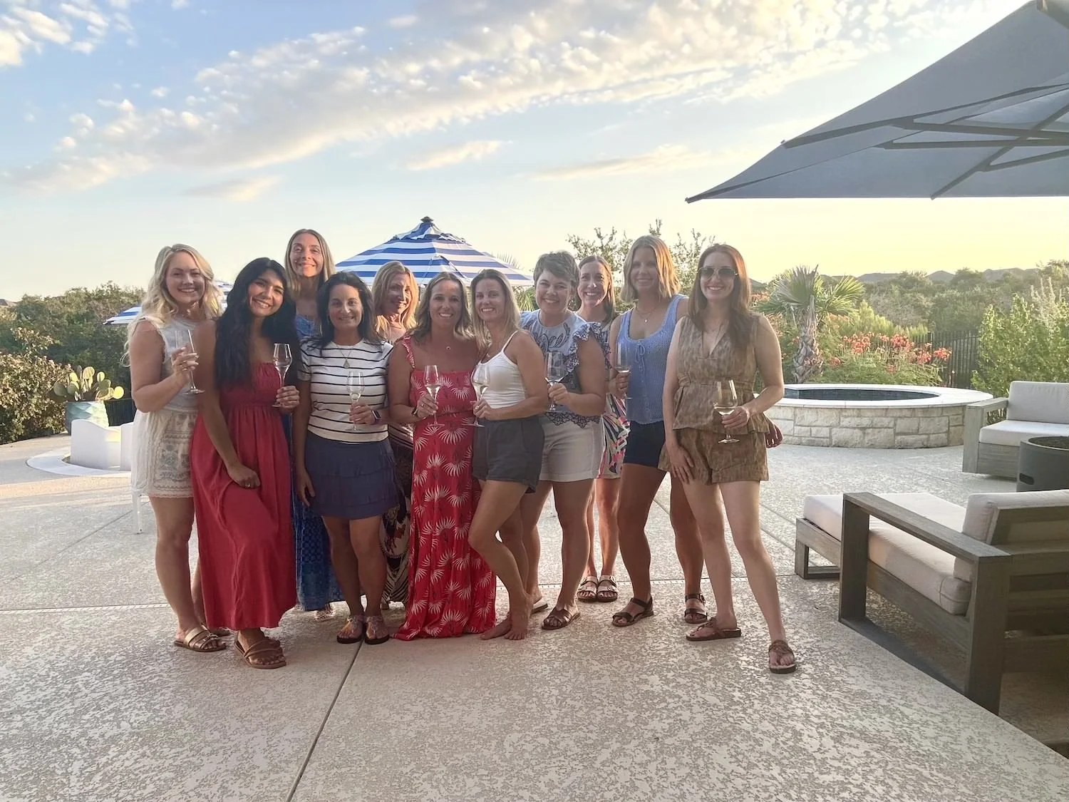 A group of women dressed in shorts and dresses standing in front of a setting sun and beautiful plants holding wine glasses