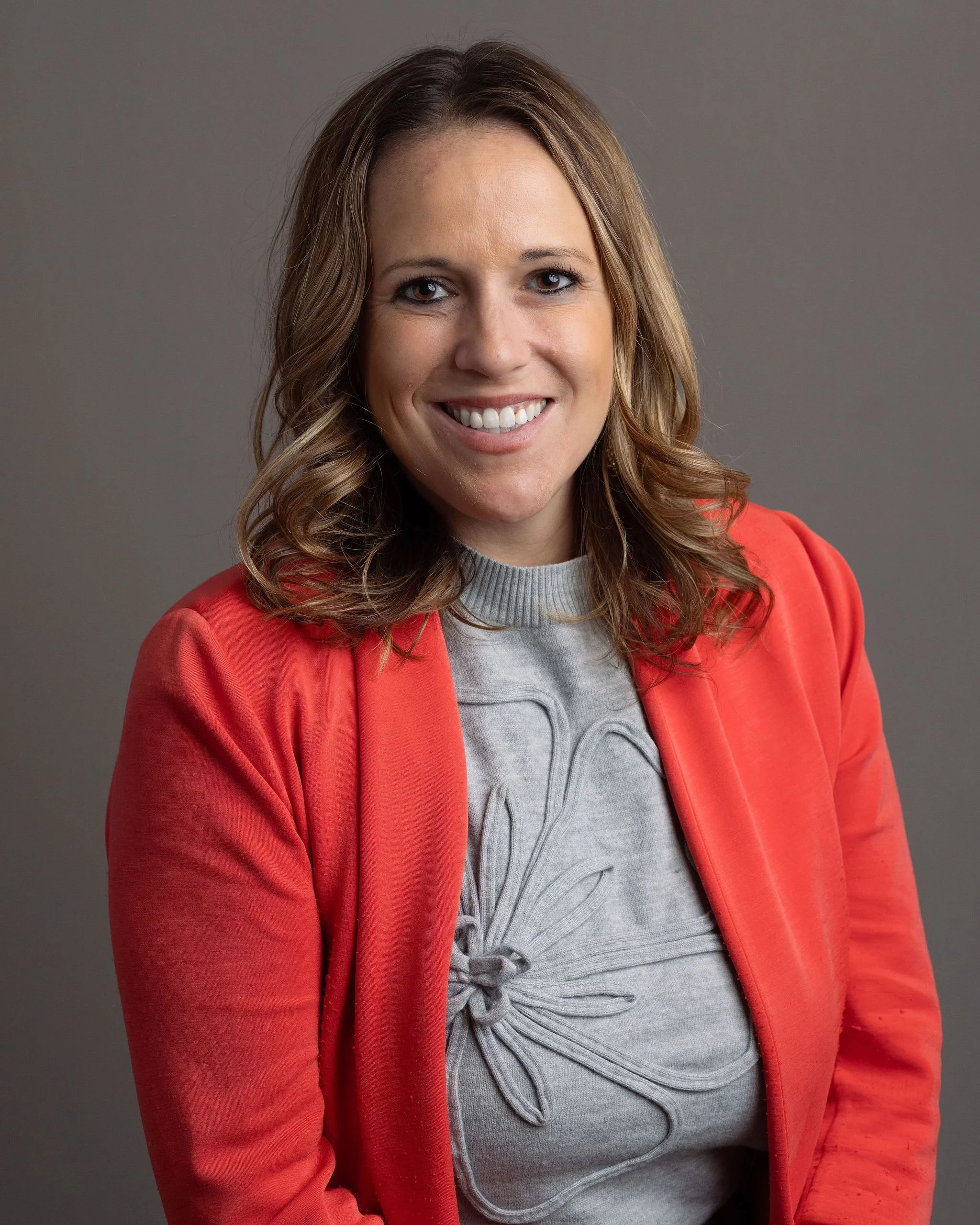 A woman with light brown, wavy hair smiling, wearing a red jacket over a gray hoodie, in front of a gray background.