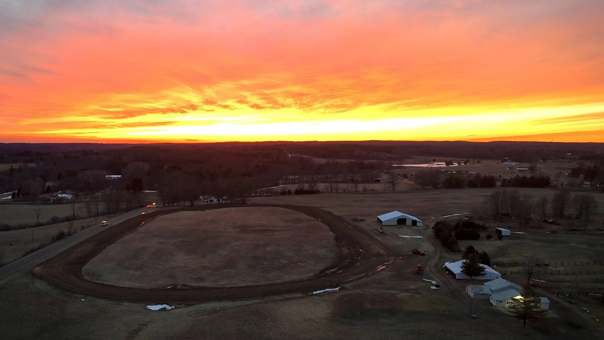 A rural landscape at sunset with an orange and yellow sky. There are several farm buildings, trees, and a winding dirt road. The grass fields are brown and there are patches of snow.