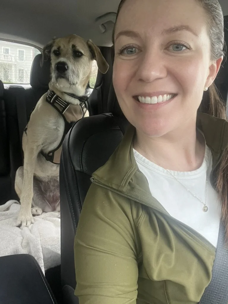 A woman smiling in the front seat of a car with a dog sitting behind her in the back seat. The dog is wearing a harness and sitting on a blanket.