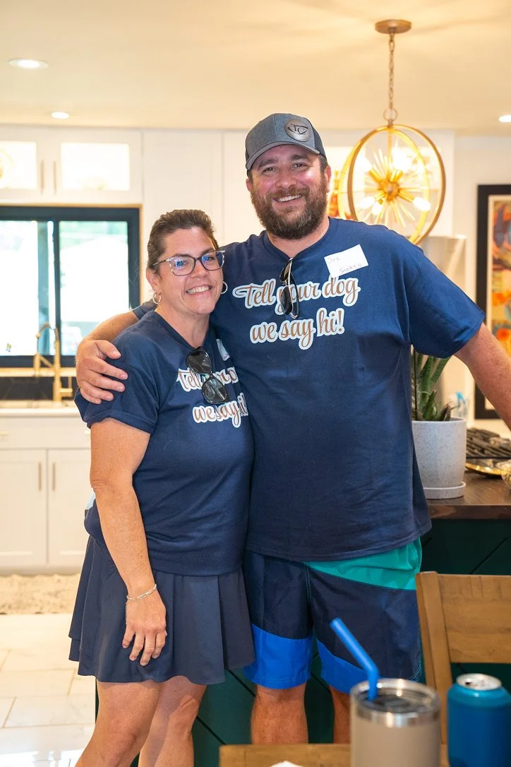 A smiling woman and man wearing matching blue T-shirts that say, 'Tell your dog we say hi!', standing in a kitchen with a modern and colorful decor, posing for the photo.