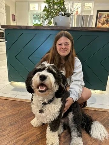 A girl with long brown hair sitting on the floor behind a large black and white dog indoors.