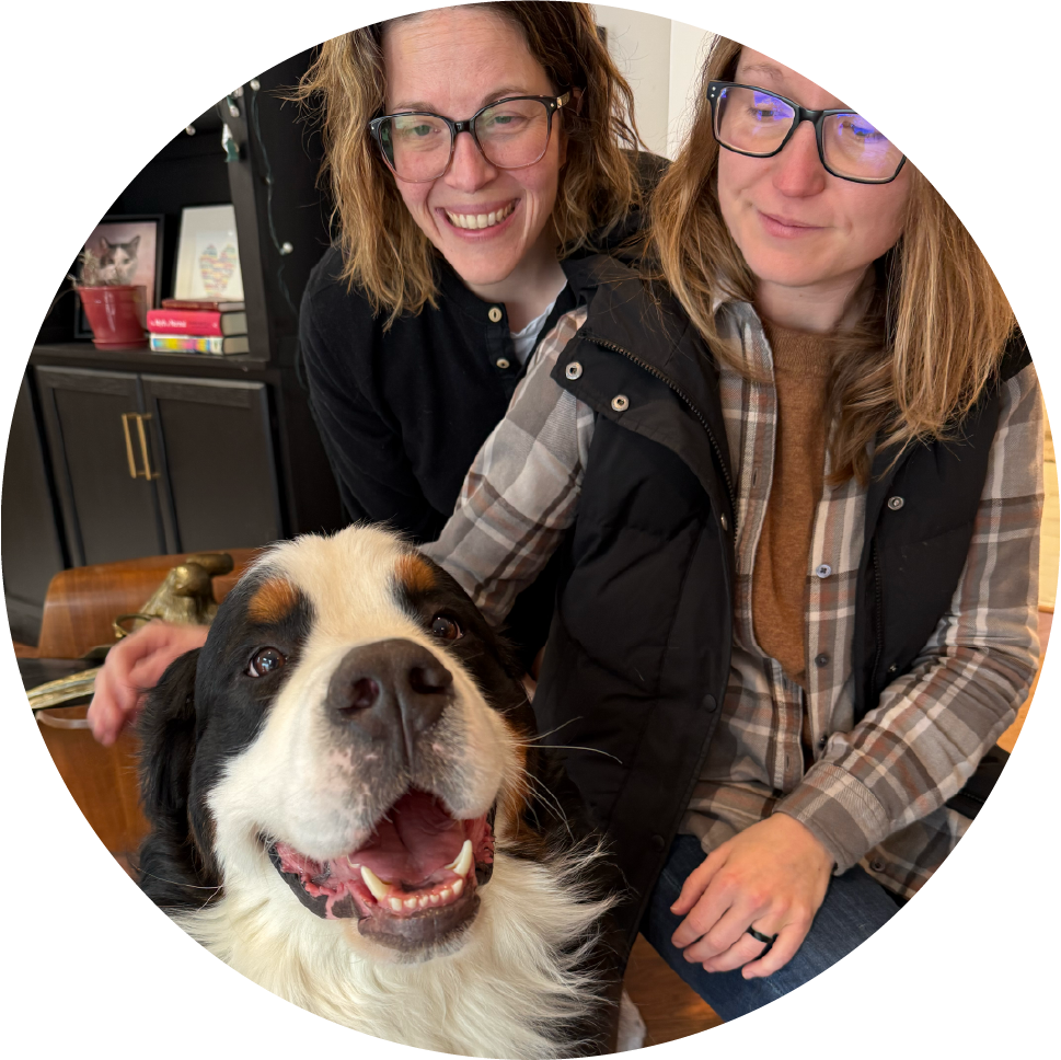 Two women smiling and petting a large black and white dog inside a house with a bookshelf and a framed picture in the background.